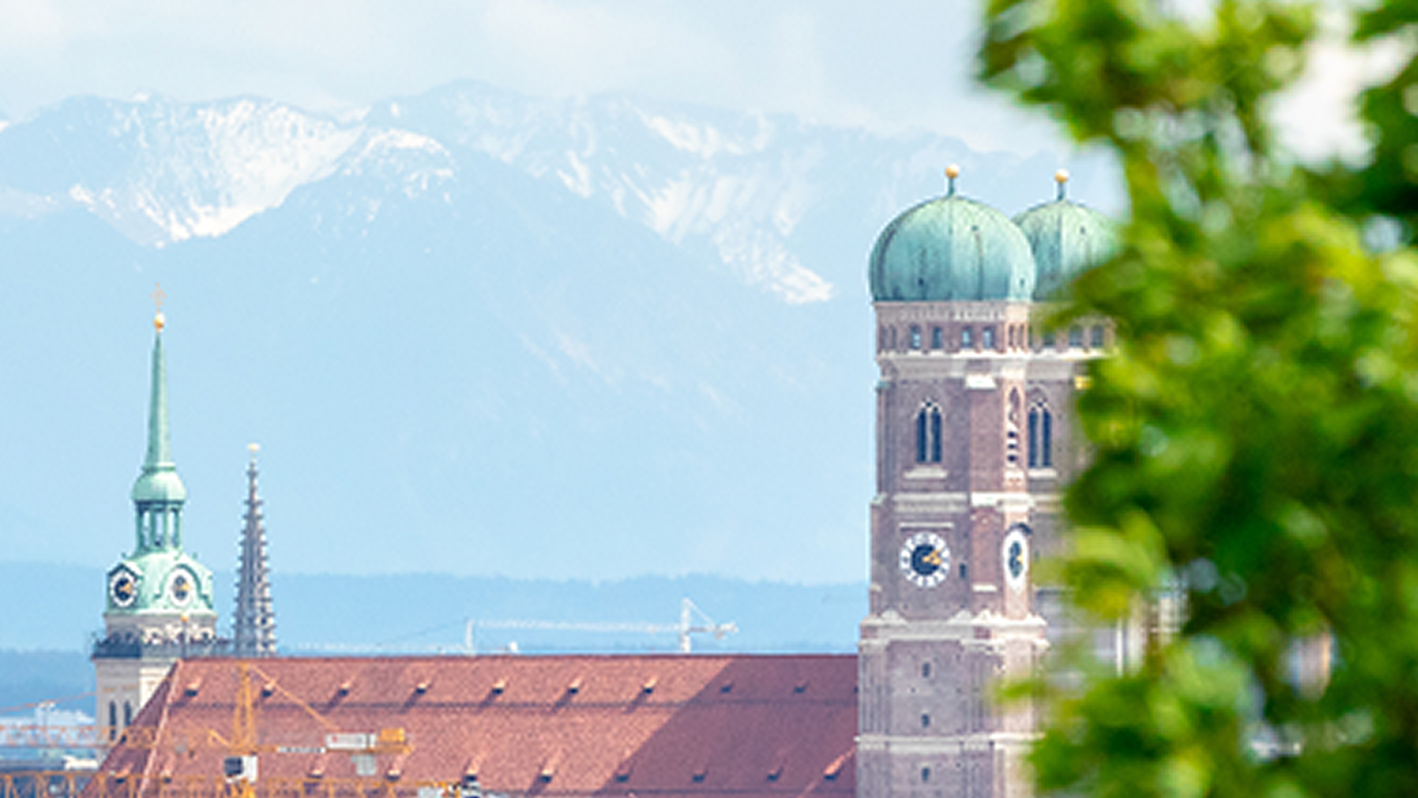 Blick auf die Frauenkirche in München mit ihren markanten Zwiebeltürmen, eingebettet in die Stadtlandschaft vor der Kulisse schneebedeckter Alpen, teils von grünen Baumblättern eingerahmt.