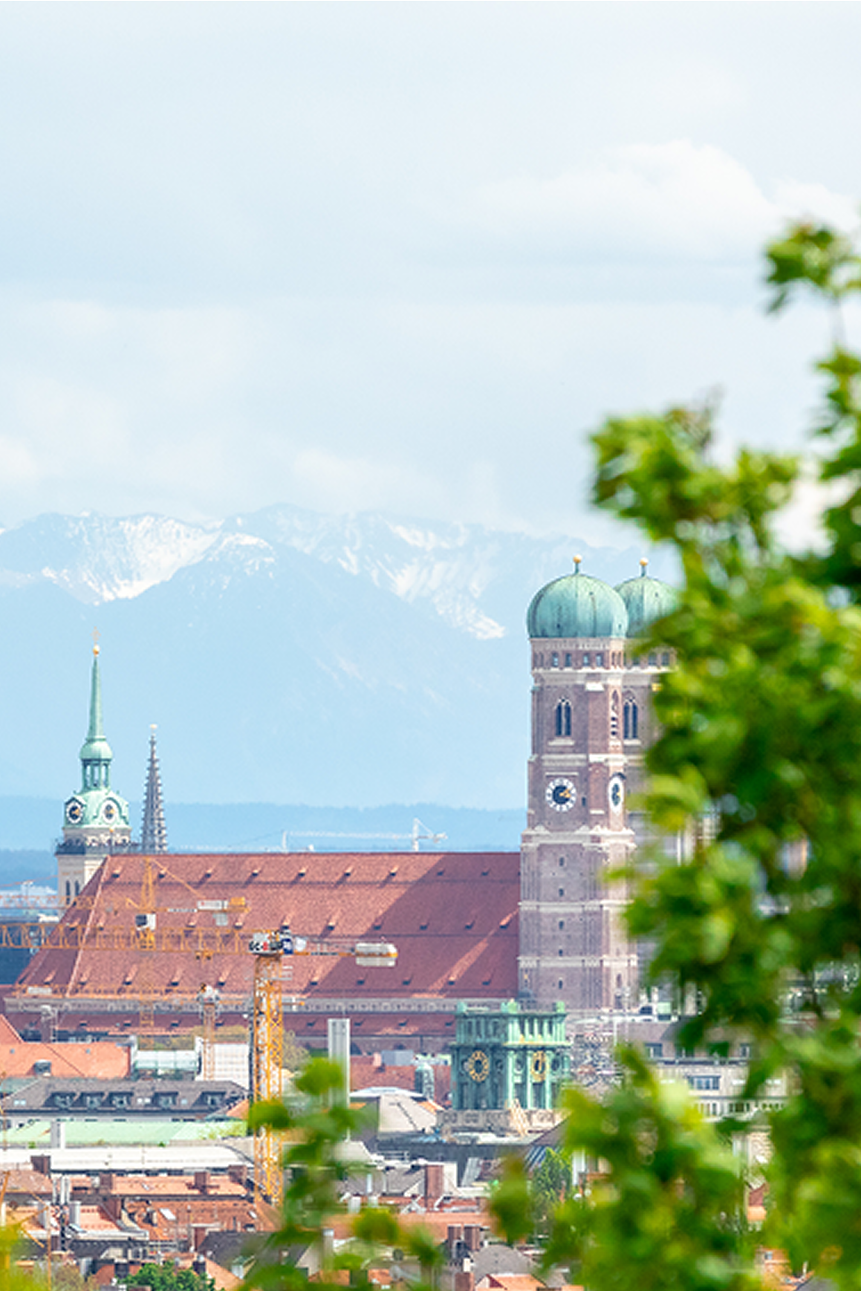 Blick auf die Frauenkirche in München mit ihren markanten Zwiebeltürmen, eingebettet in die Stadtlandschaft vor der Kulisse schneebedeckter Alpen, teils von grünen Baumblättern eingerahmt.