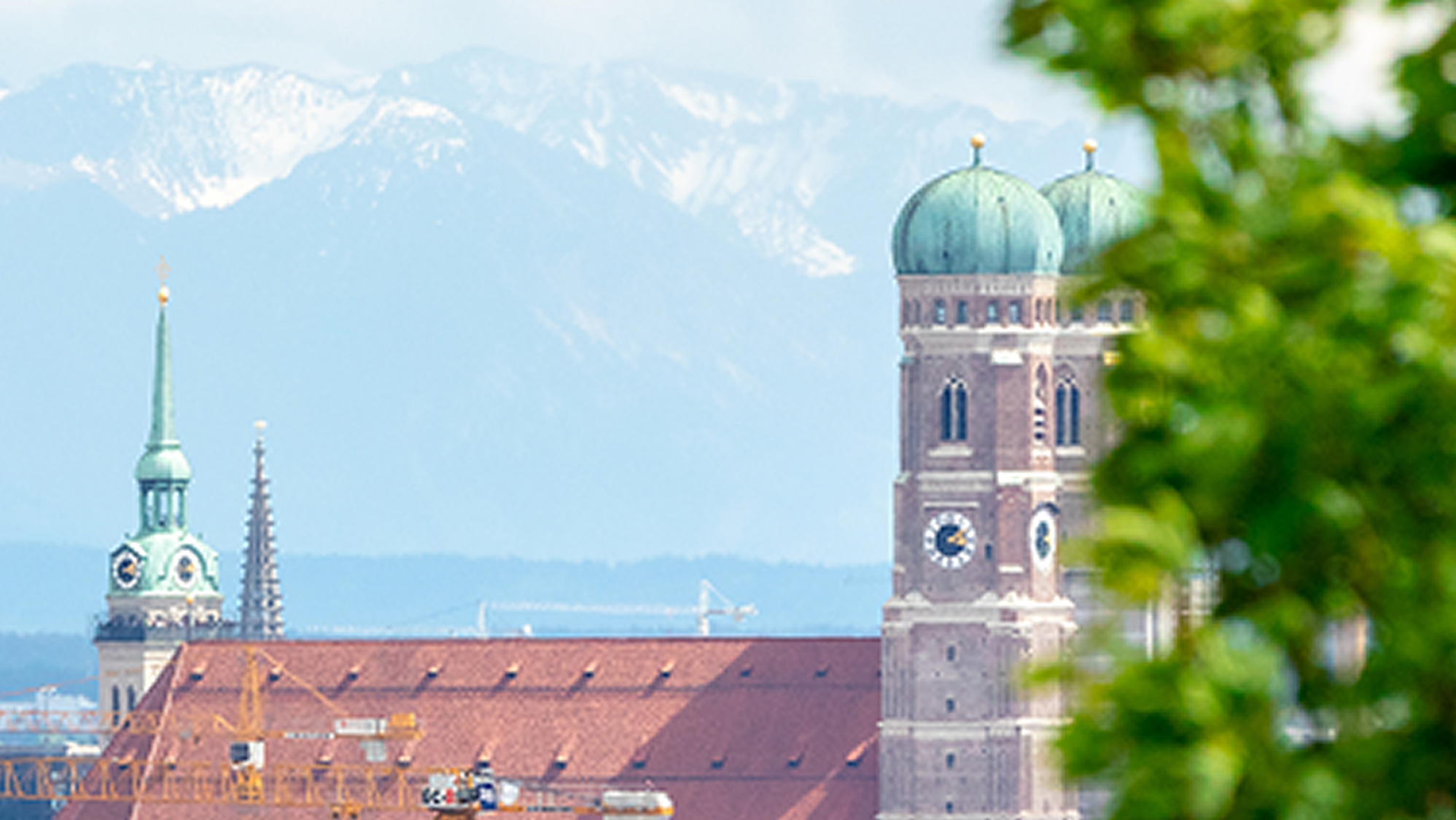 Blick auf die Frauenkirche in München mit ihren markanten Zwiebeltürmen, eingebettet in die Stadtlandschaft vor der Kulisse schneebedeckter Alpen, teils von grünen Baumblättern eingerahmt.