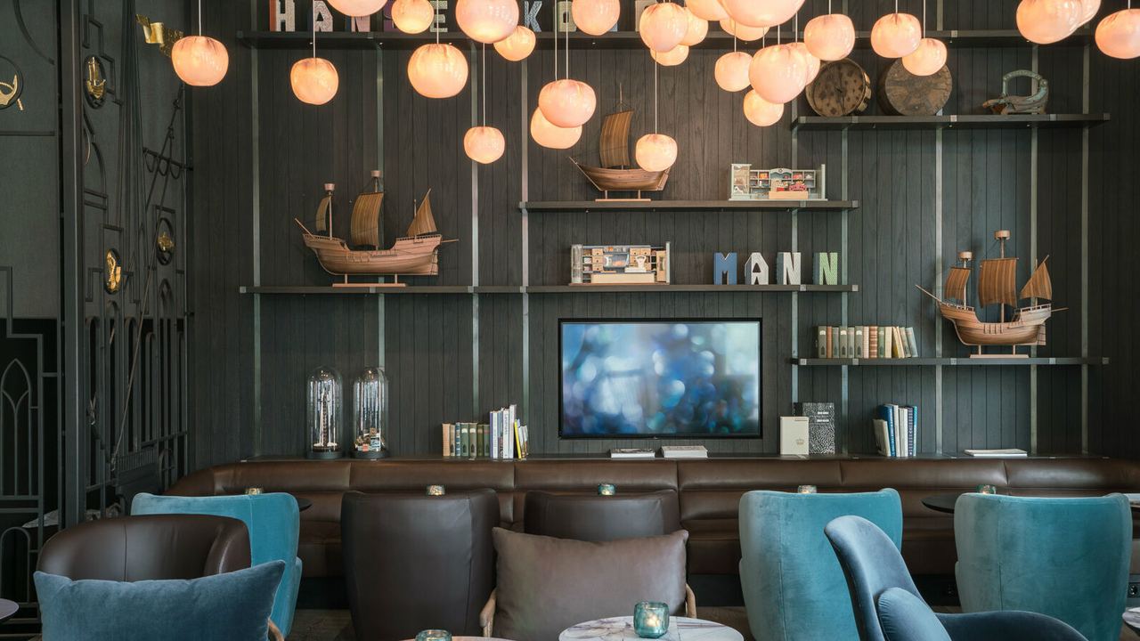 Gemütliche Lobby mit dunkelblauen Sesseln, runden Tischen und weicher Beleuchtung durch viele hängende Lampen, ergänzt durch maritime Deko und Bücherregale für entspannte Aufenthalte.