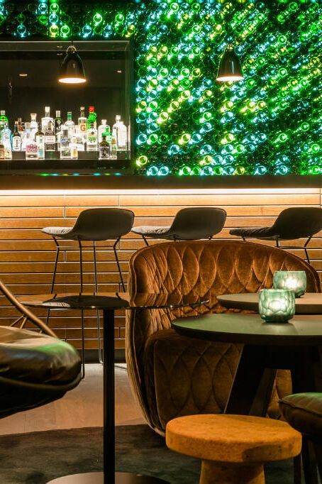 Gemütliche Hotelbar mit modernem Design, Leder-Sitzmöbeln und Hängekorb. Im Hintergrund leuchtet eine Wand aus grünen Flaschen, die für eine besondere Atmosphäre sorgt. Die Bar zeigt eine Auswahl an Spirituosen, ideal für entspannte Abende.