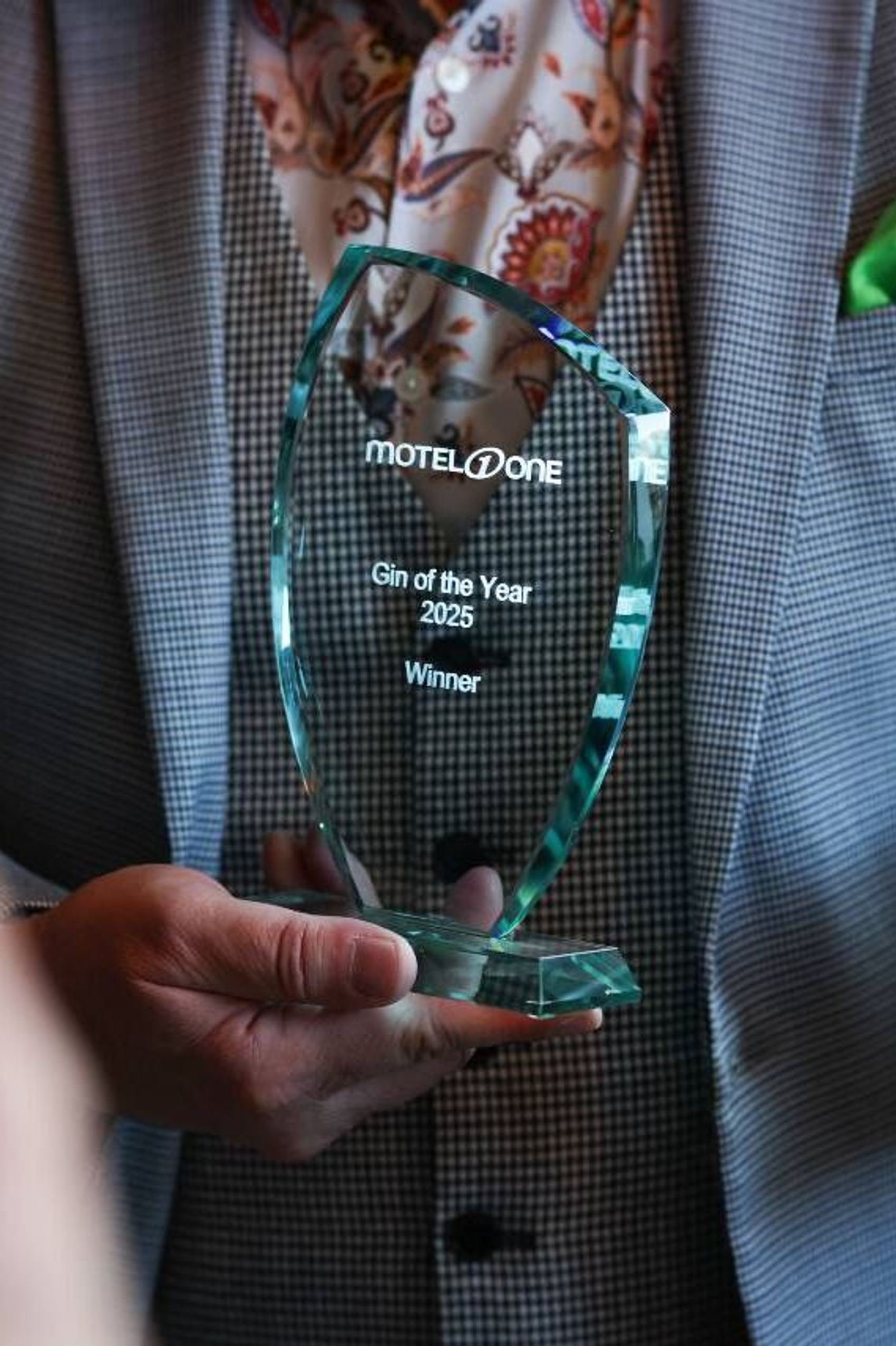 Person in Anzug hält eine Auszeichnung mit der Aufschrift „Motel One Gift of the Year 2025 Winner“ und eine dekorative Flasche, was Erfolg und Anerkennung symbolisiert.