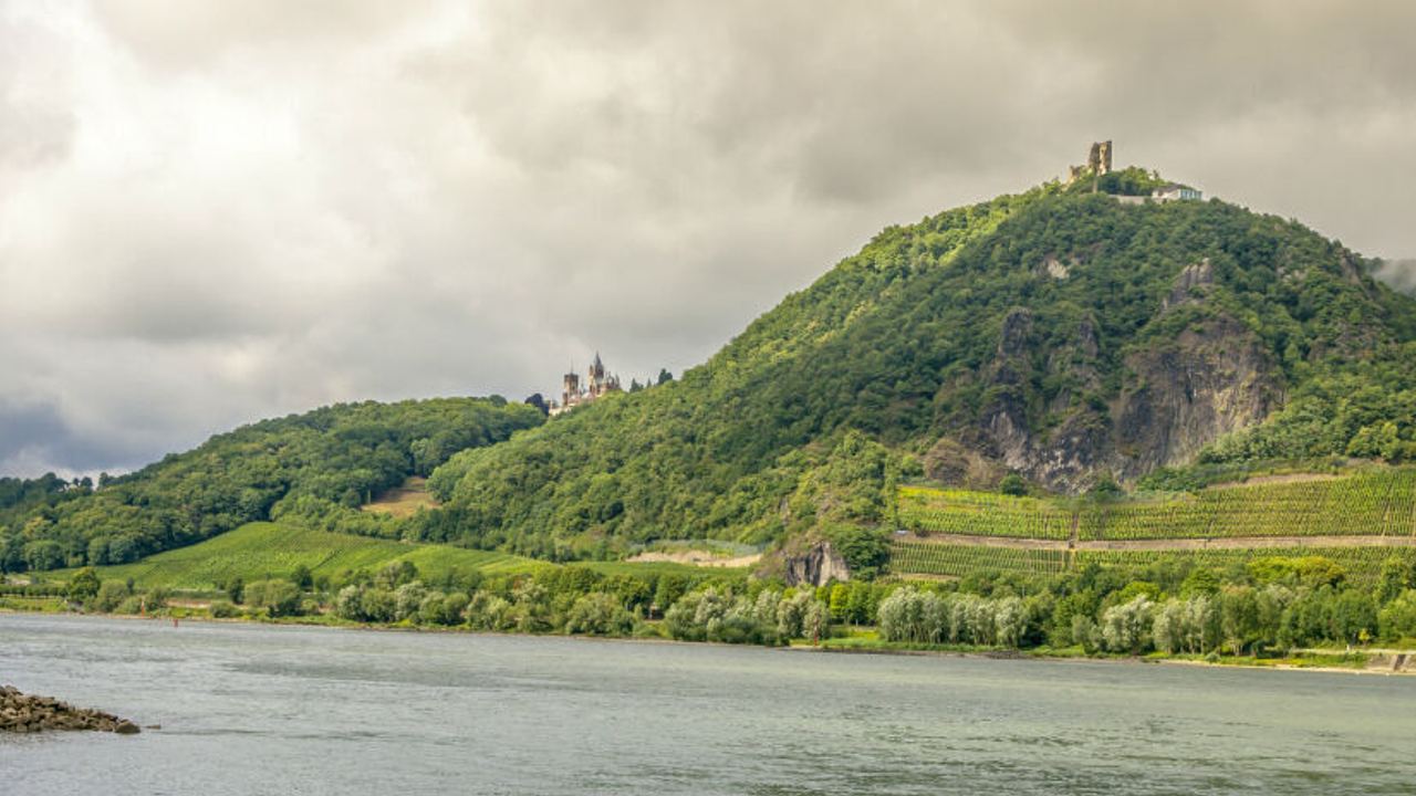 Flusslandschaft mit steinigem Ufer, dahinter grüne bewaldete Hügel mit Weinbergen und zwei historischen Burgruinen unter wolkigem Himmel.