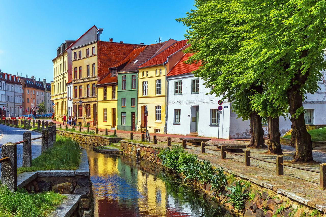 Bunte, historische Fachwerkhäuser entlang eines ruhigen Kanals unter klarem, blauem Himmel, mit grünen Bäumen und einem gepflasterten Uferweg, der zum entspannten Spazieren einlädt.