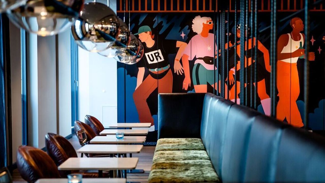 Moderne Bar mit gemütlicher Sitzbank, Ledersesseln und weißen Tischen; farbenfrohe Wandmalerei zeigt stilisierte Figuren in Bewegung, schafft lebendige, künstlerische Atmosphäre für gesellige Abende.