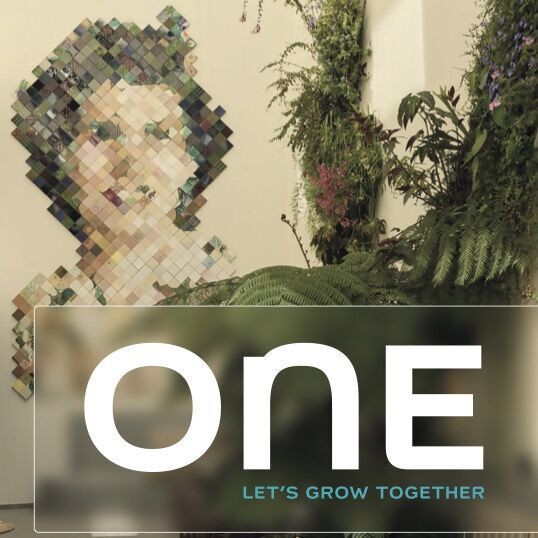 Moderne Lobby mit grünen Pflanzen und stilvoller Sitzgelegenheit. Wandkunst zeigt ein pixeliertes Gesicht, symbolisiert Zusammenhalt. Schriftzug „ONE – Let’s Grow Together“ unterstreicht Gemeinschaft und Wachstum.