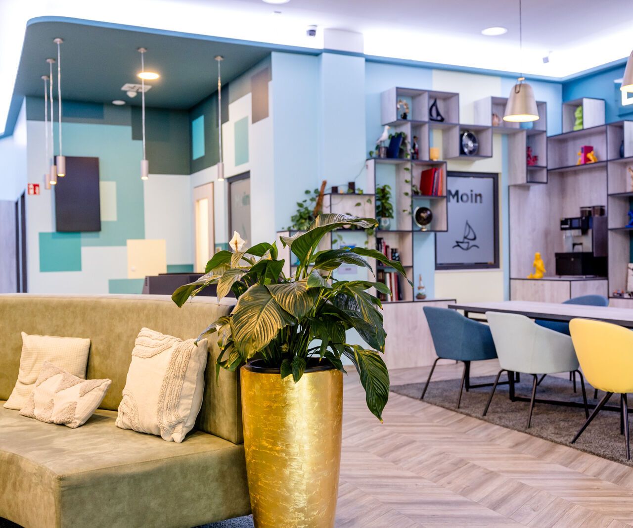 Heller und moderner Aufenthaltsraum mit gemütlicher Sitzecke, Farbakzenten, großen Pflanzen und mehreren Sitzplätzen. Bücherregale an der Wand schaffen eine entspannte, einladende Atmosphäre für Gäste zum Lesen oder Treffen.