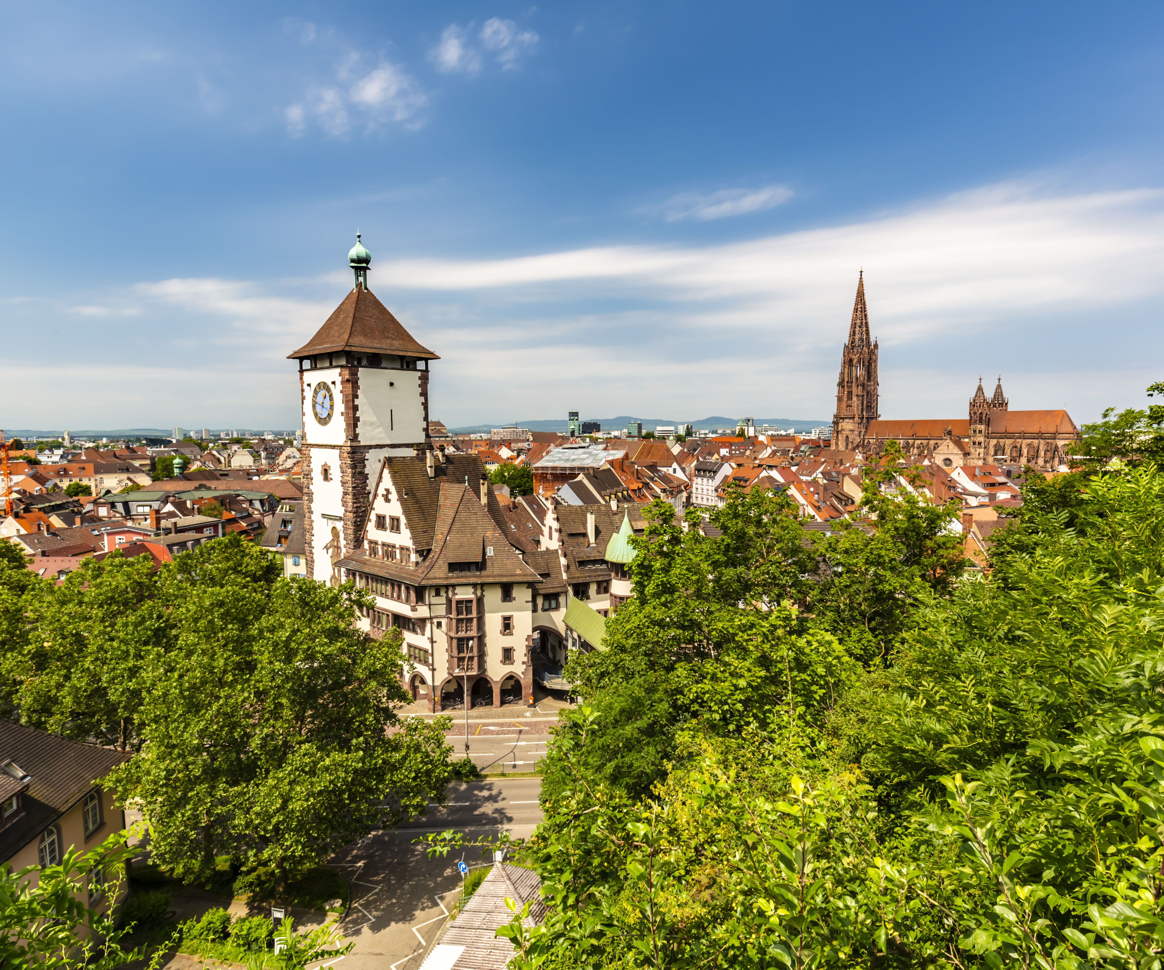 Blick auf die Altstadt mit historischem Uhrturm, umgeben von grünen Bäumen, unter blauem Himmel, im Hintergrund die Kathedrale einer europäischen Stadt.