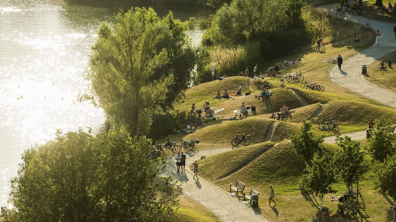 Grüne Parklandschaft am Wasser mit sanften Hügeln und Spazierwegen, auf denen Menschen entspannen, spazieren gehen und Fahrrad fahren, umgeben von Bäumen und natürlichem Licht.