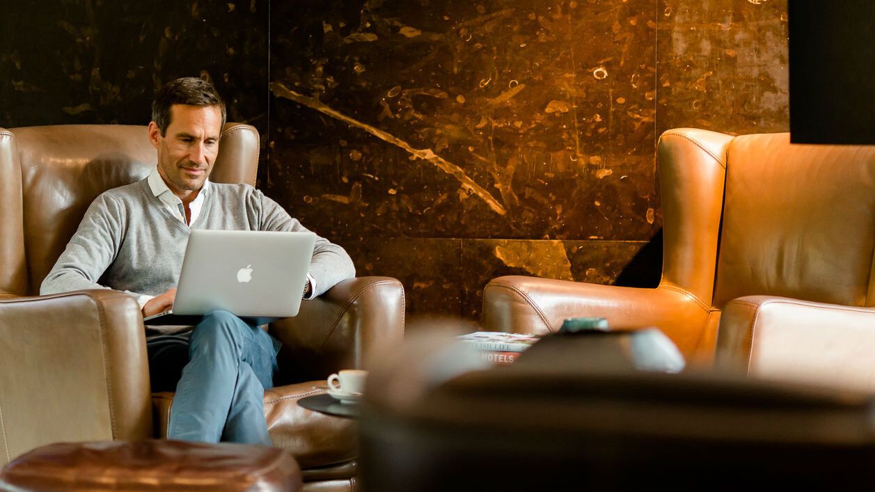 Ein Mann arbeitet entspannt mit einem Laptop in einer gemütlichen Lounge mit Lederstühlen und warmem, einladendem Ambiente.
