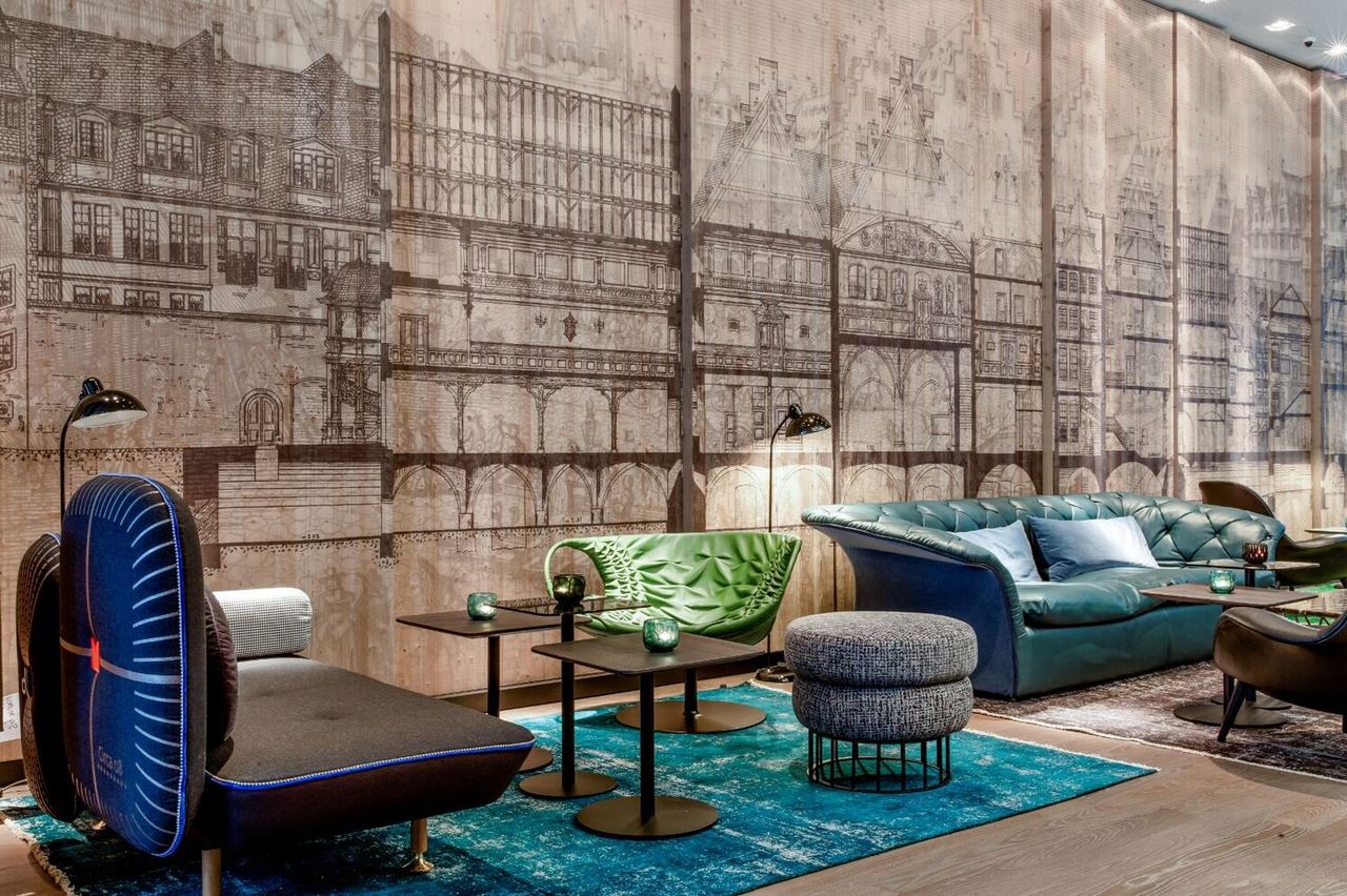 Motel One Lounge mit stilvollen Sitzmöbeln in Blau und Grün, runden schwarzen Tischen und einer Wandgestaltung mit historischen Gebäudezeichnungen