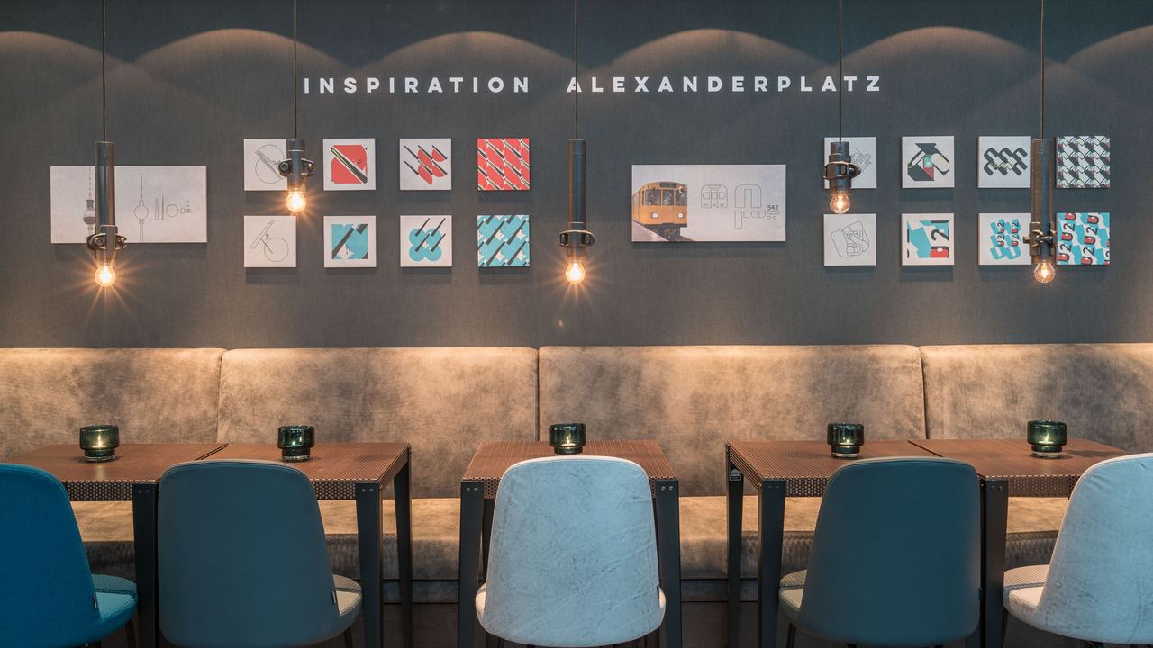 Hotel Berlin Alexanderplatz Motel One Essbereich mit mehreren Tischen und Stühlen sowie Schriftzug Inspiration Alexanderplatz an der Wand