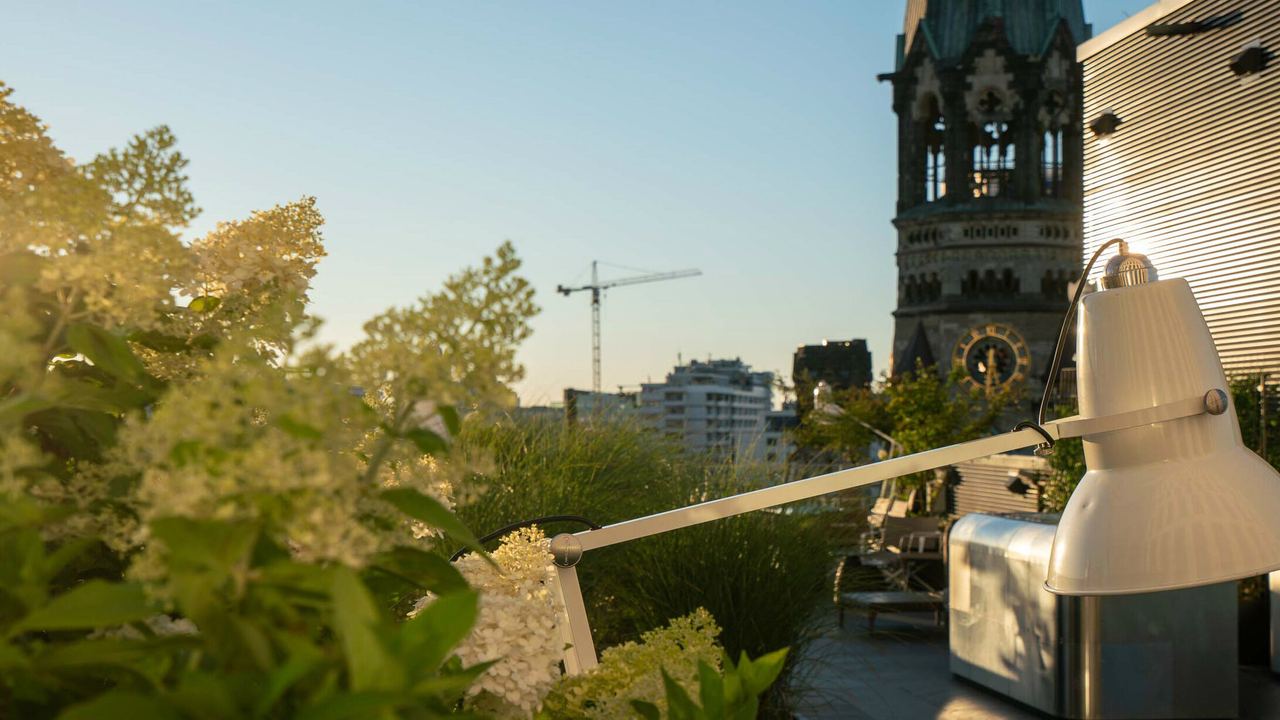Blick von einer begrünten Dachterrasse bei Sonnenuntergang auf die Kaiser-Wilhelm-Gedächtniskirche in Berlin, mit moderner Beleuchtung und entspannter Atmosphäre.