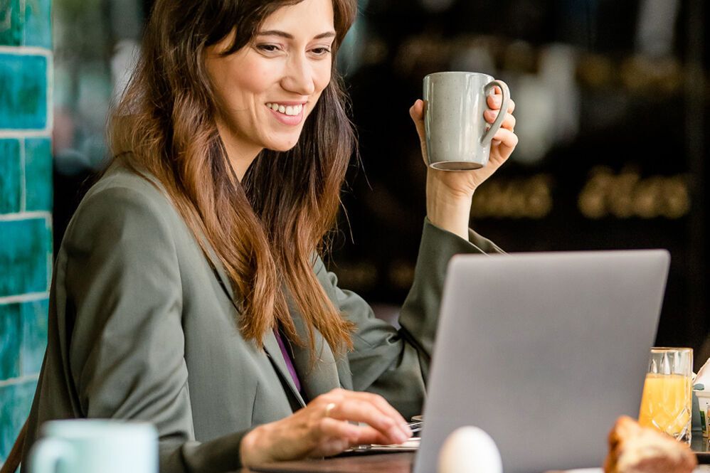 Junge Frau arbeitet entspannt mit Laptop in gemütlichem Café, hält Tasse in der Hand, genießt Frühstück mit frischem Gebäck und Saft.