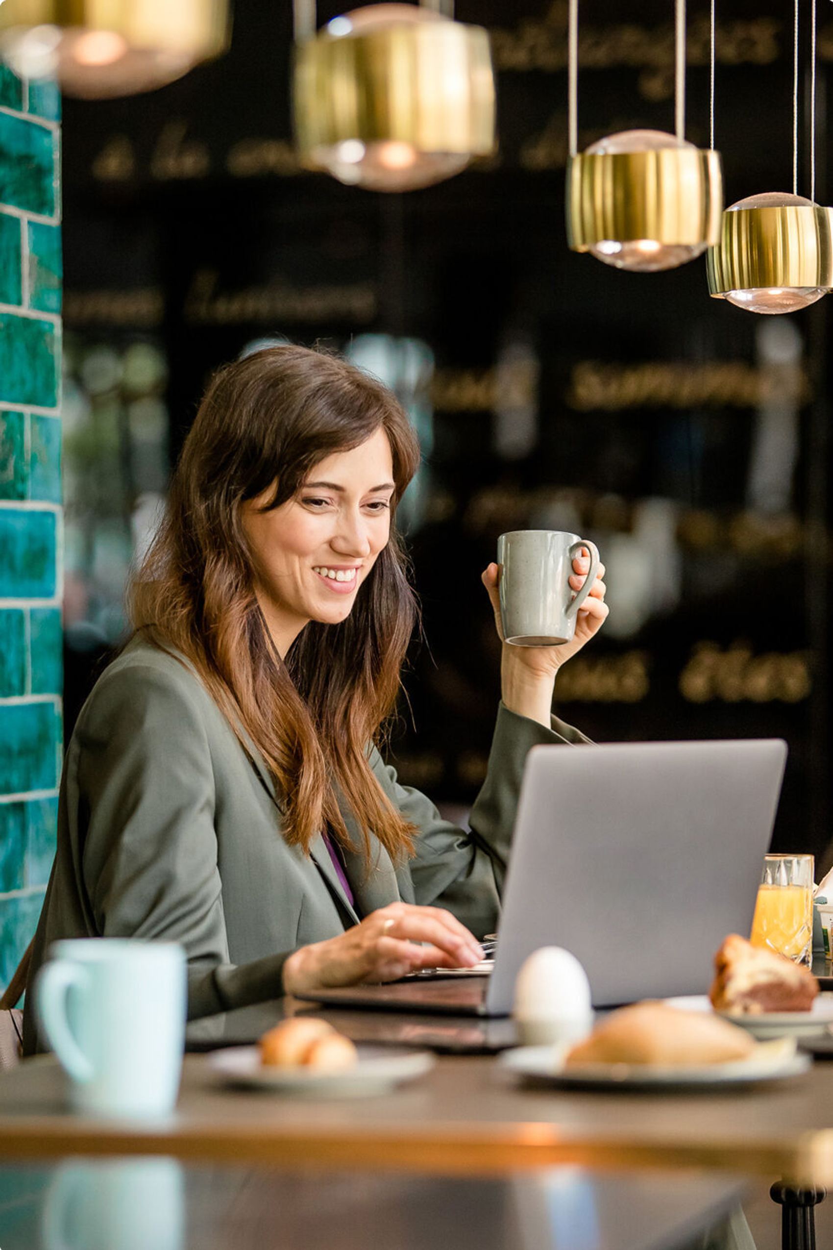 Junge Frau arbeitet entspannt mit Laptop in gemütlichem Café, hält Tasse in der Hand, genießt Frühstück mit frischem Gebäck und Saft.