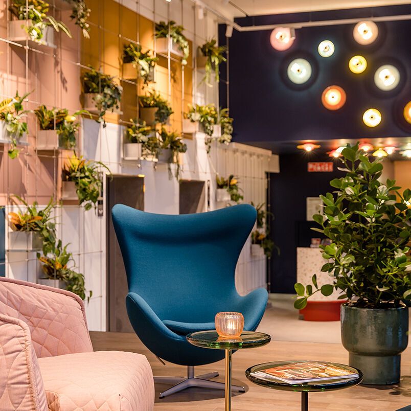 Motel One Lounge mit modernem blauen Sessel, rosa Sofa und grünen Pflanzen. Warme, dekorative Wandbeleuchtung 