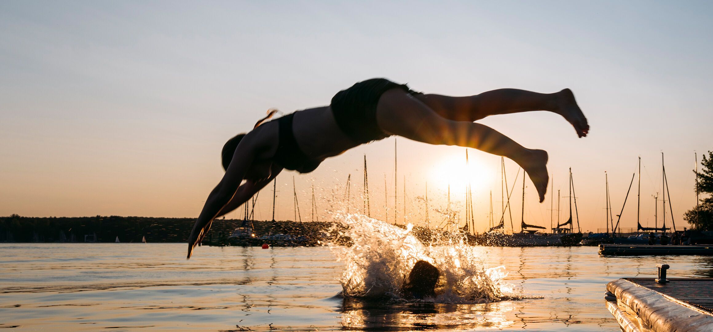 Person bei Sonnenuntergang in einem See, die ins Wasser springt, mit Segelbooten im Hintergrund und ruhiger Wasseroberfläche.