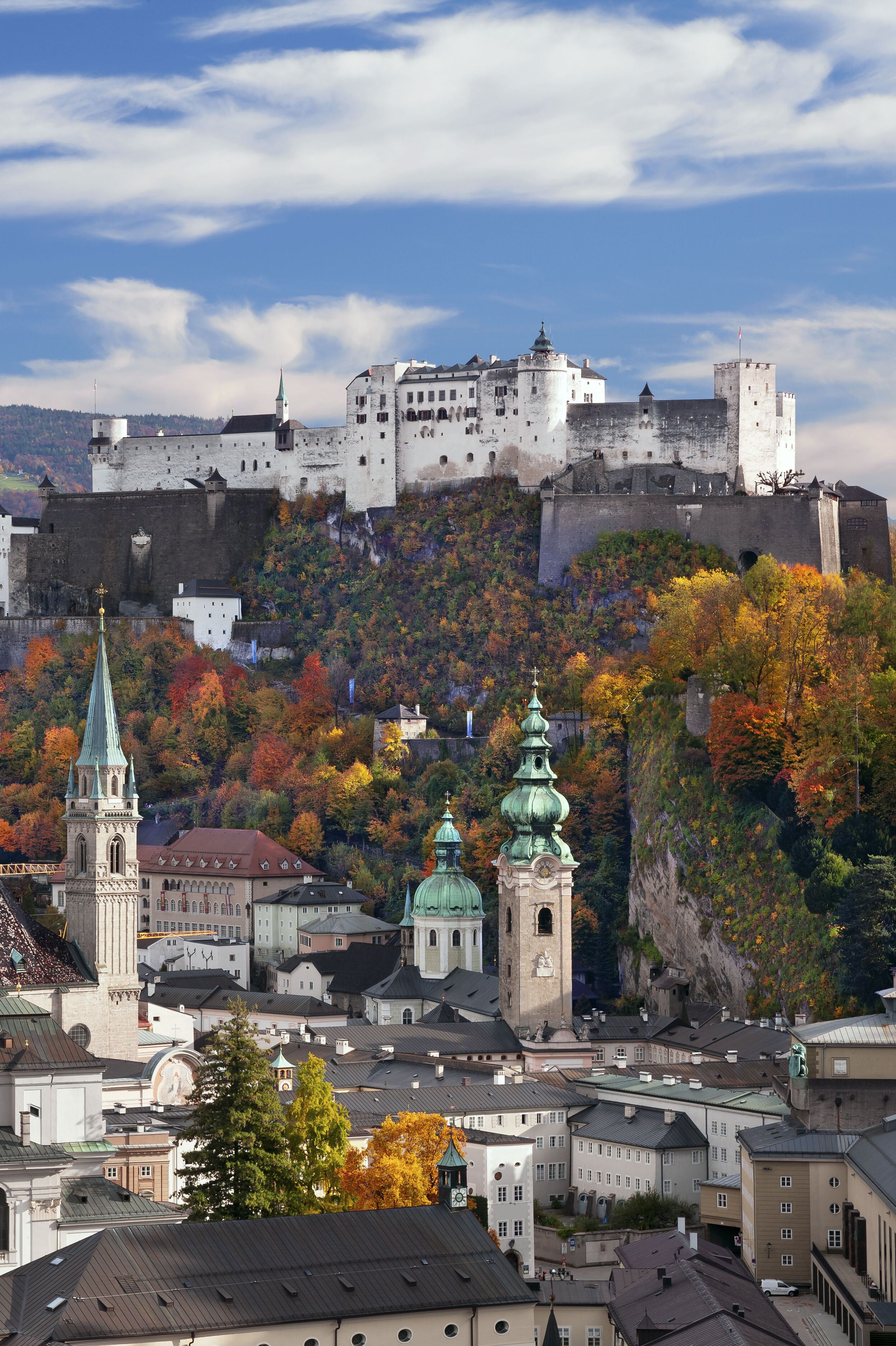 Blick auf die historische Altstadt von Salzburg mit zahlreichen Kirchen und der Festung Hohensalzburg auf einem bewaldeten Hügel, umgeben von herbstlich bunt gefärbten Bäumen unter klarem Himmel. Blick auf die historische Altstadt von Salzburg mit zahlreichen Kirchen und der Festung Hohensalzburg auf einem bewaldeten Hügel, umgeben von herbstlich bunt gefärbten Bäumen unter klarem Himmel.