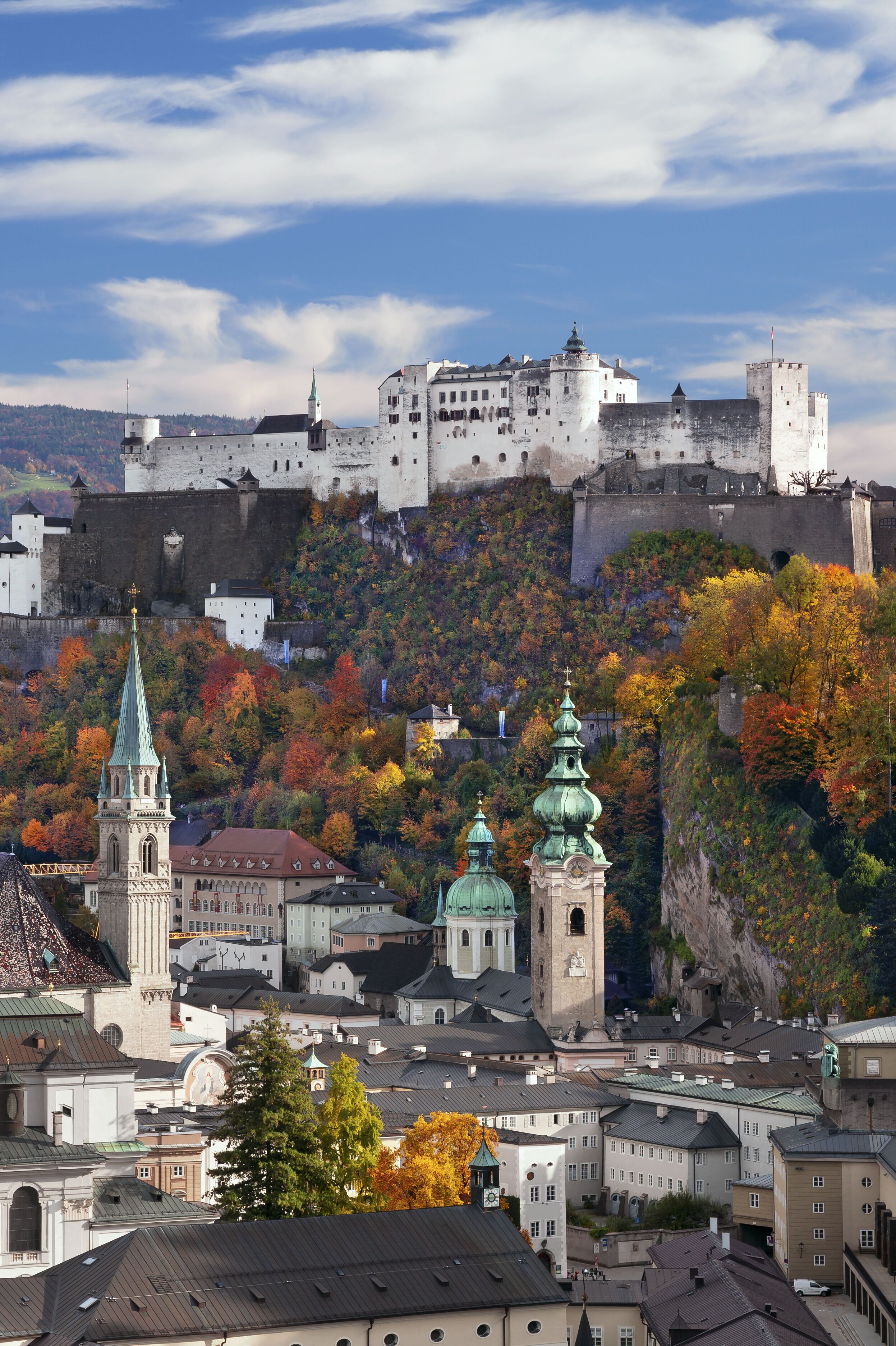 Blick auf die historische Altstadt von Salzburg mit zahlreichen Kirchen und der Festung Hohensalzburg auf einem bewaldeten Hügel, umgeben von herbstlich bunt gefärbten Bäumen unter klarem Himmel.
