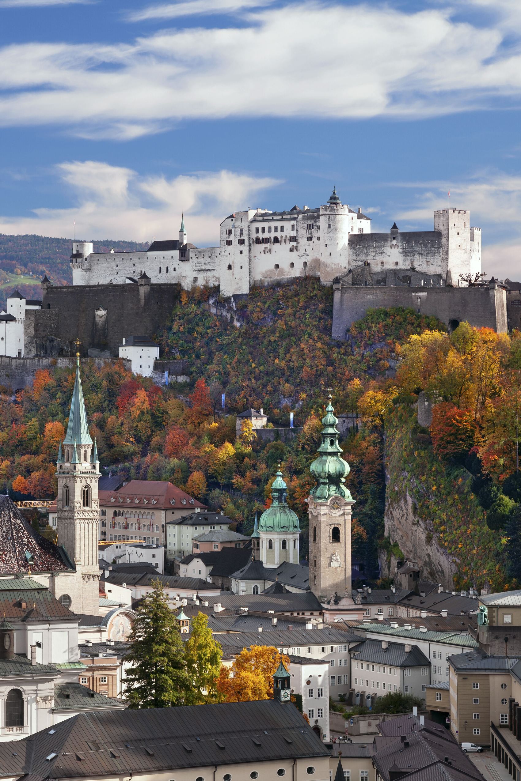 Blick auf die historische Altstadt von Salzburg mit zahlreichen Kirchen und der Festung Hohensalzburg auf einem bewaldeten Hügel, umgeben von herbstlich bunt gefärbten Bäumen unter klarem Himmel.