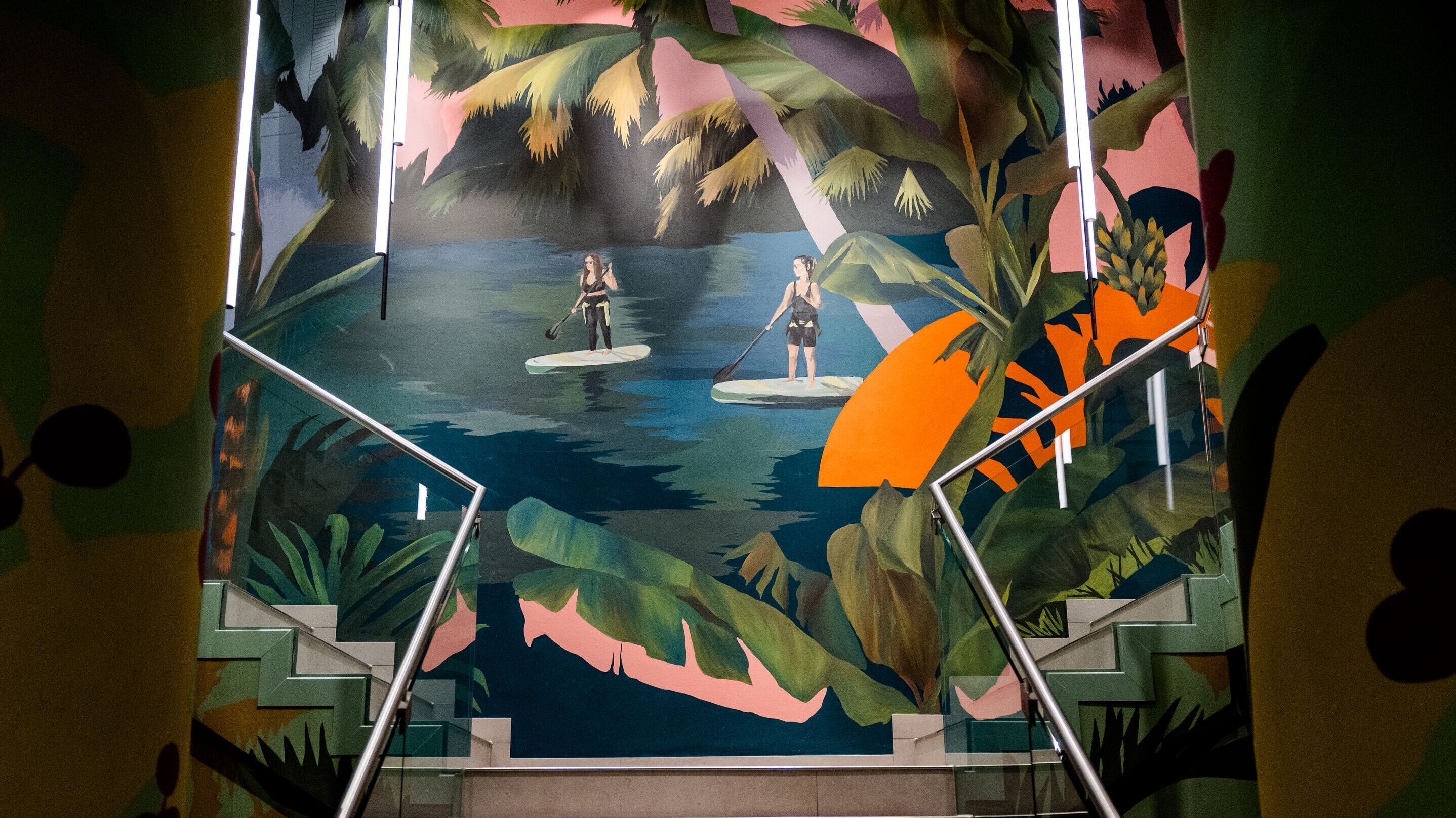 Bunte Wandmalerei in einem Treppenhaus zeigt zwei Personen auf Paddleboards in einem tropischen Gewässer, umgeben von Palmen und großen Blättern, vermittelt Urlaubsfeeling und Entspannung.