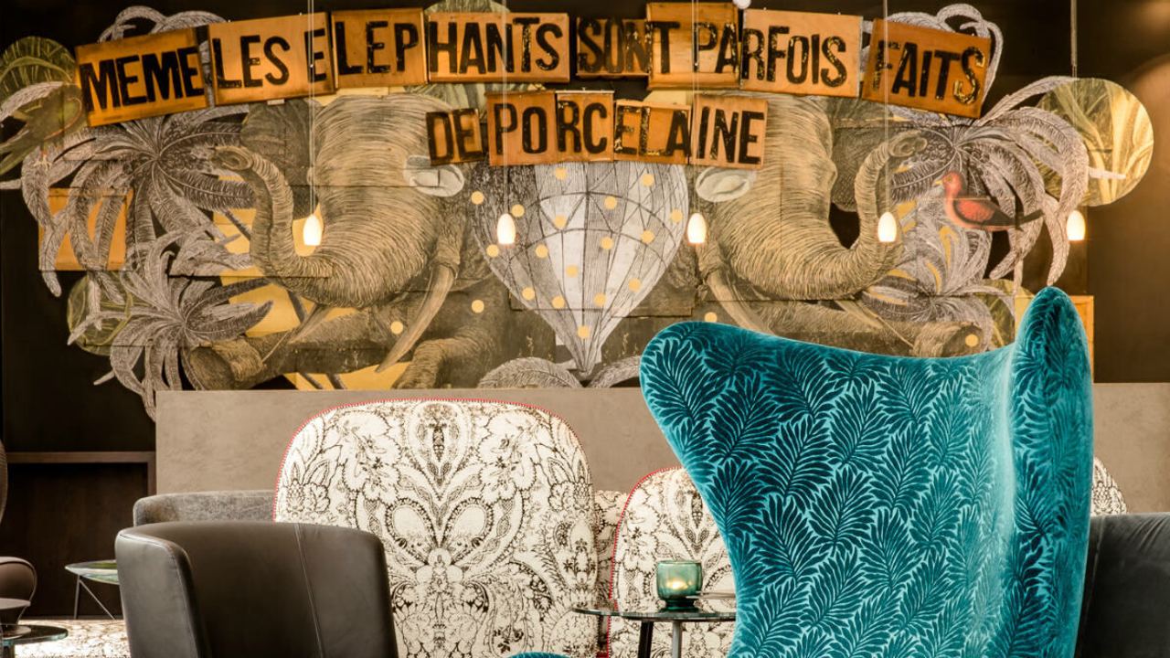 Motel One Lounge-Bereich mit modernem, türkisfarbenem Ohrensessel, gemusterten und schwarzen Sesseln vor einer Wand mit künstlerischer Elefanten-Illustration und französischem Schriftzug.