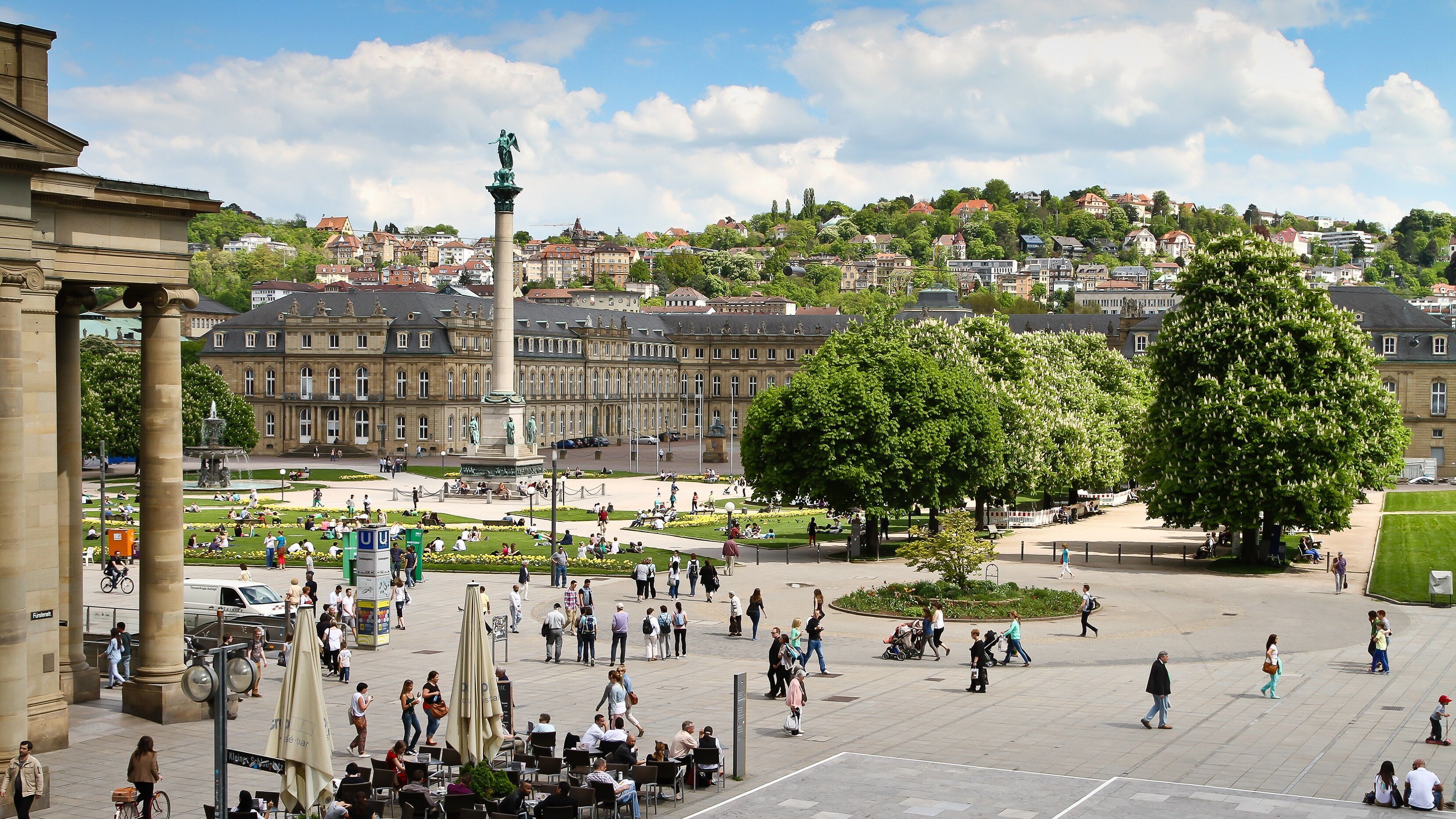 Blick auf einen belebten, sonnigen Platz mit einer hohen Säule in der Mitte, von historischen Gebäuden umgeben, umgeben von grünen Bäumen und Hügeln im Hintergrund, viele Menschen genießen die Atmosphäre und entspannen im Freien.