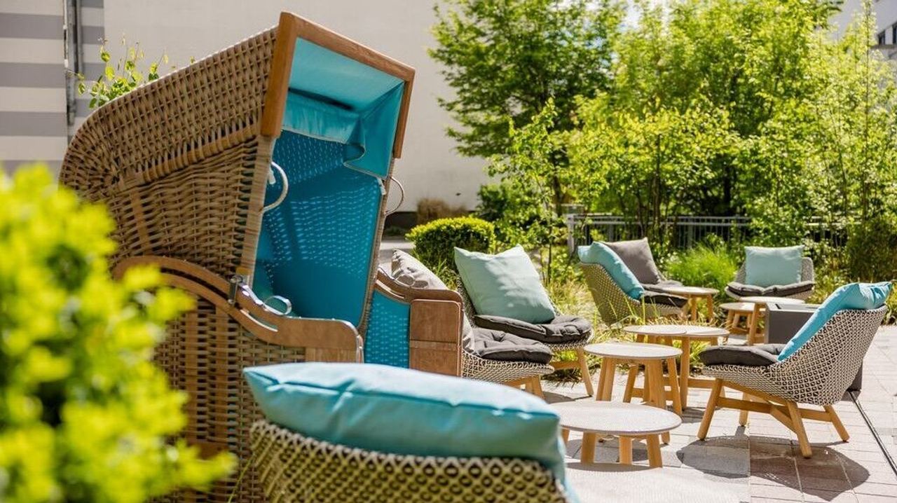 Gemütliche Außenlounge mit Korbsessel und gepolsterten Stühlen, umgeben von grünen Pflanzen, ideal für entspannte Stunden im Freien.