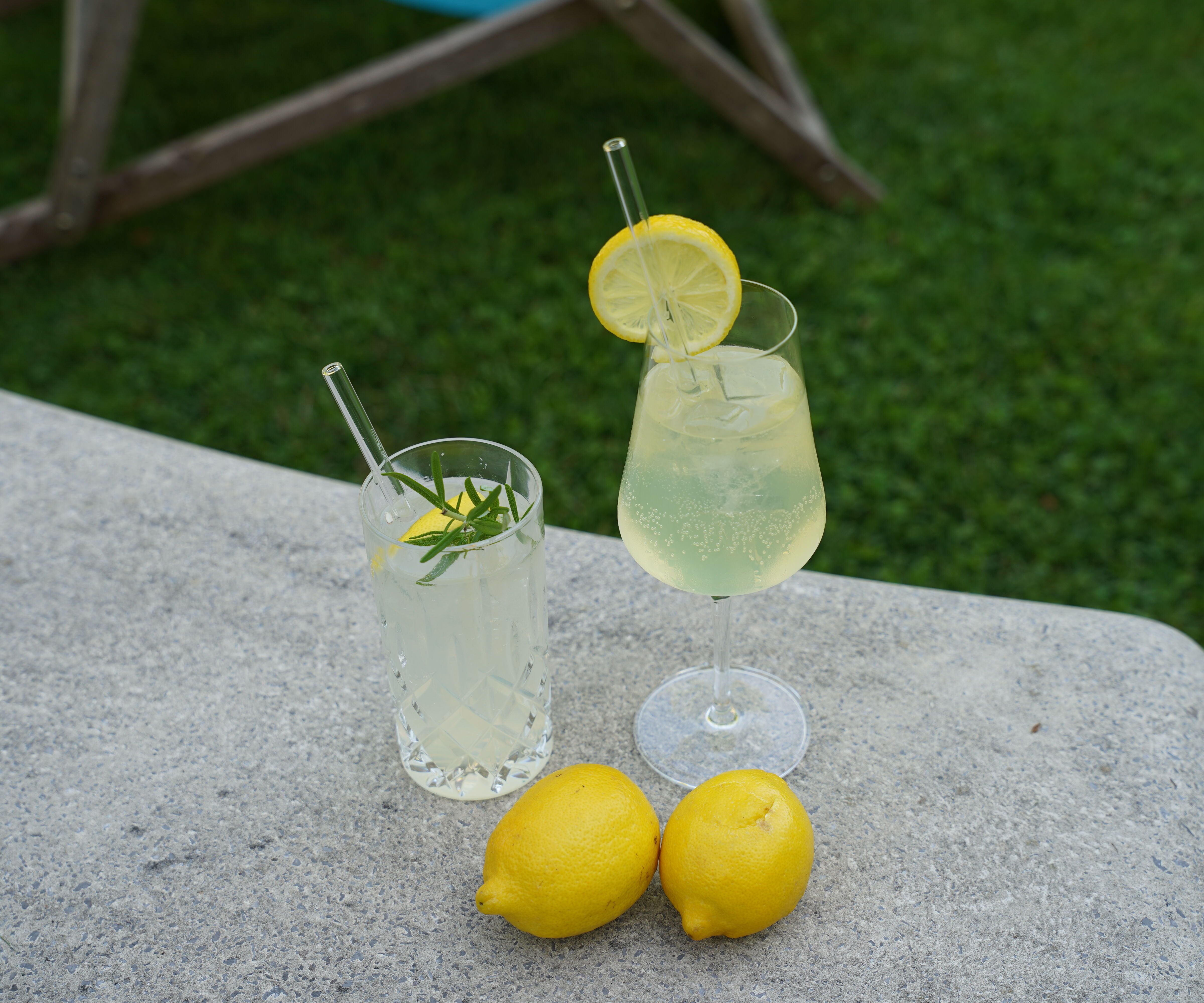 Zwei erfrischende Zitronengetränke mit Eis und Minze stehen auf einem grauen Steintisch, daneben liegen zwei frische Zitronen, im Hintergrund grüner Rasen.