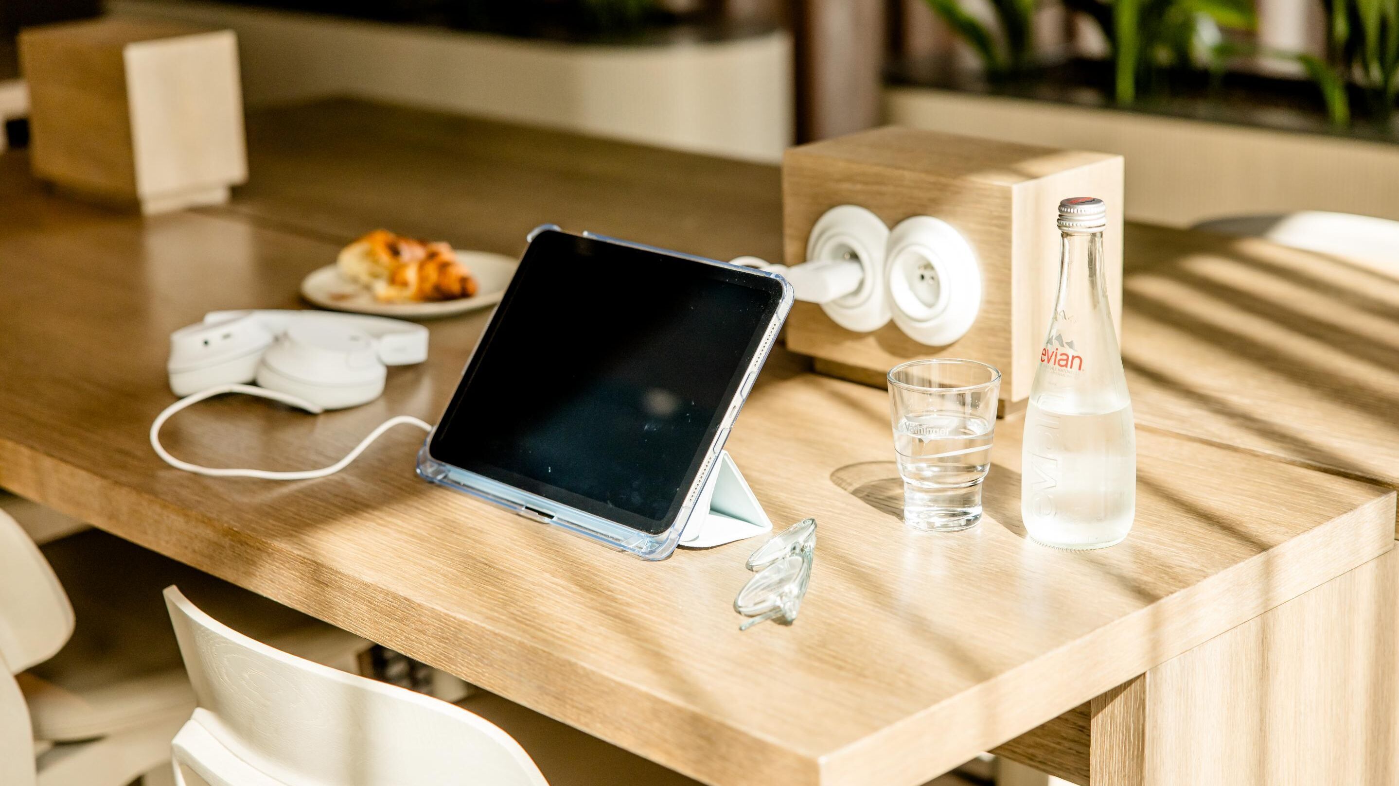Moderner Arbeitsplatz in Motel One Lounge mit Tablet, das an einer Steckdose geladen wird, daneben eine Flasche Wasser und ein Glas. 