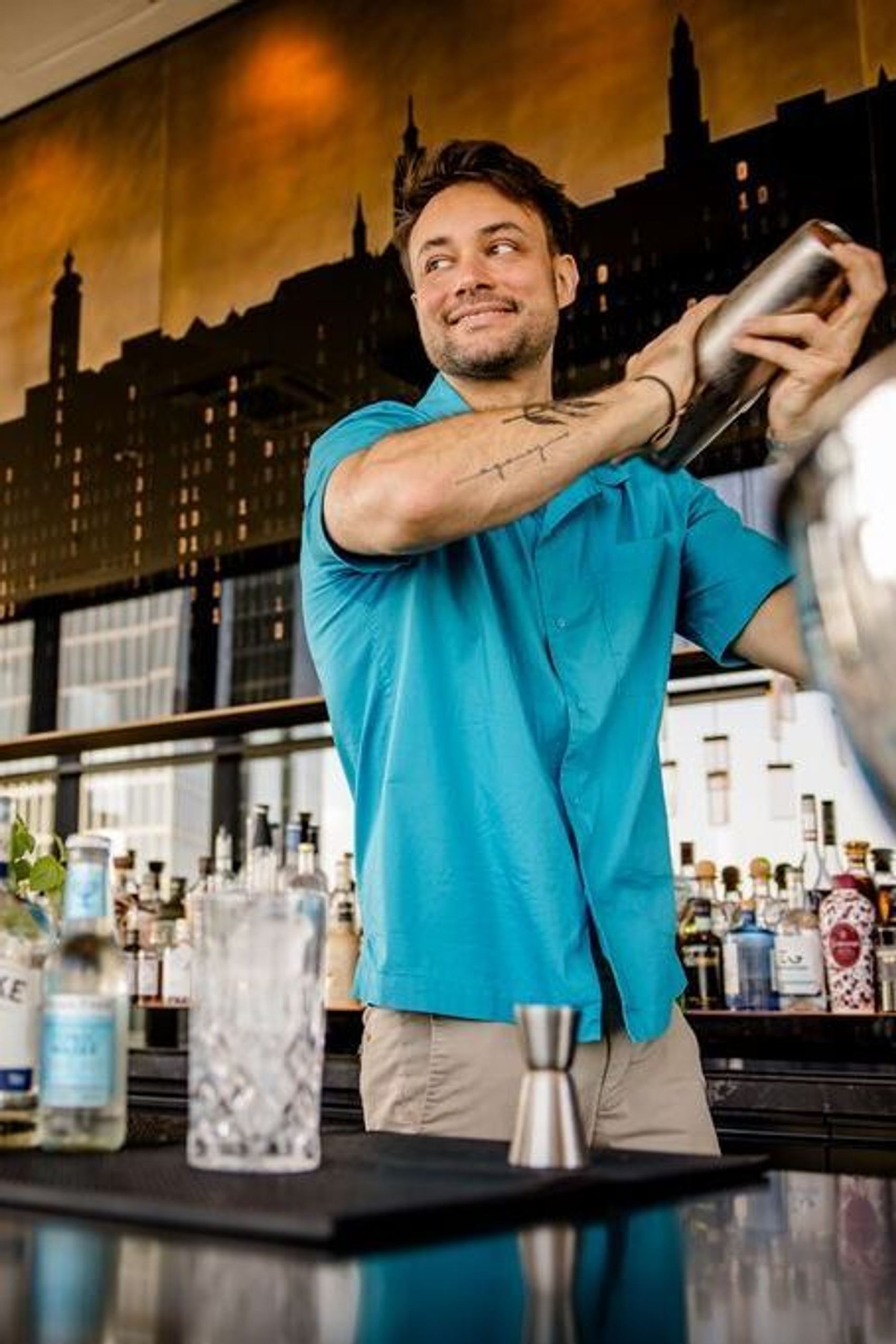 Ein Barkeeper in einem modernen Hotelbar-Umfeld mixt einen Cocktail und lächelt dabei, umgeben von verschiedenen Spirituosen und Glaswaren.