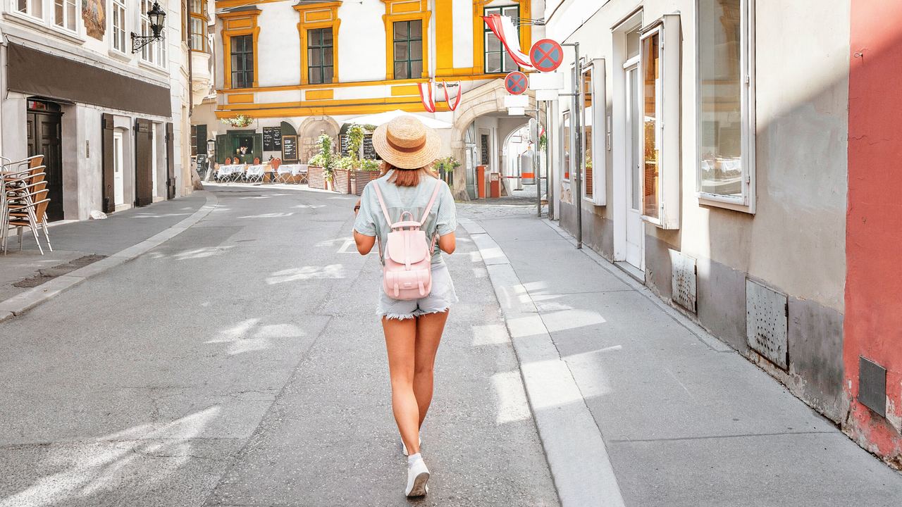 Eine Frau mit sommerlichem Outfit und Hut schlendert durch eine historische, sonnige Altstadtgasse mit bunten, gut erhaltenen Gebäuden und gemütlicher Atmosphäre.