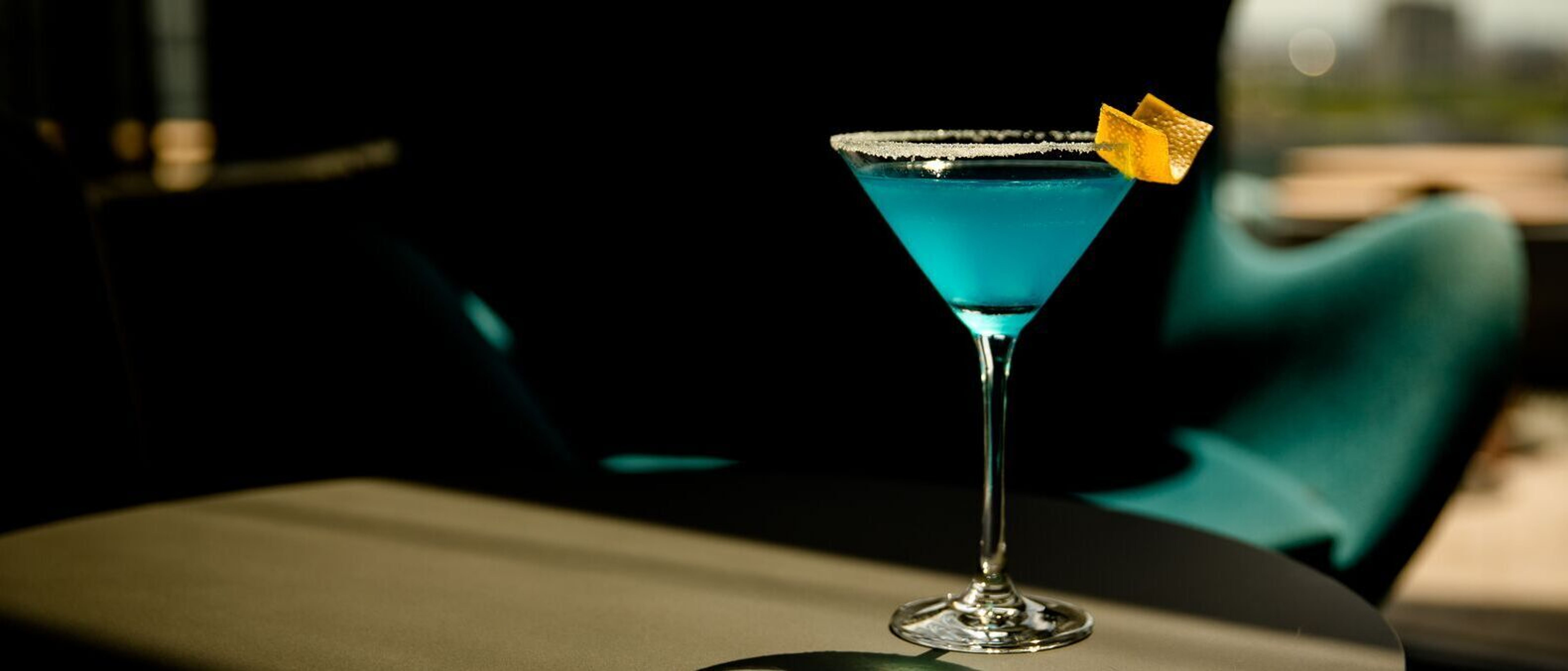 Ein blauer Cocktail in einem Martiniglas mit Zuckerrand und Orangenzeste steht auf einem runden Tisch, im Hintergrund ein gemütlicher Sitzbereich mit großen Fenstern und Tageslicht.