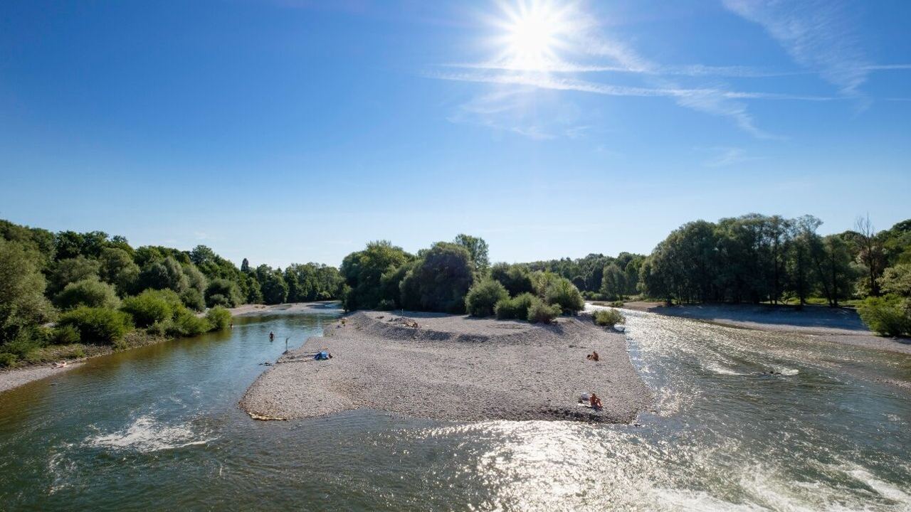 Fluss mit einer kleinen Kiesinsel in der Mitte, umgeben von grünem Wald, unter strahlend blauem Himmel mit heller Sonne. Einige Menschen entspannen auf der Insel.
