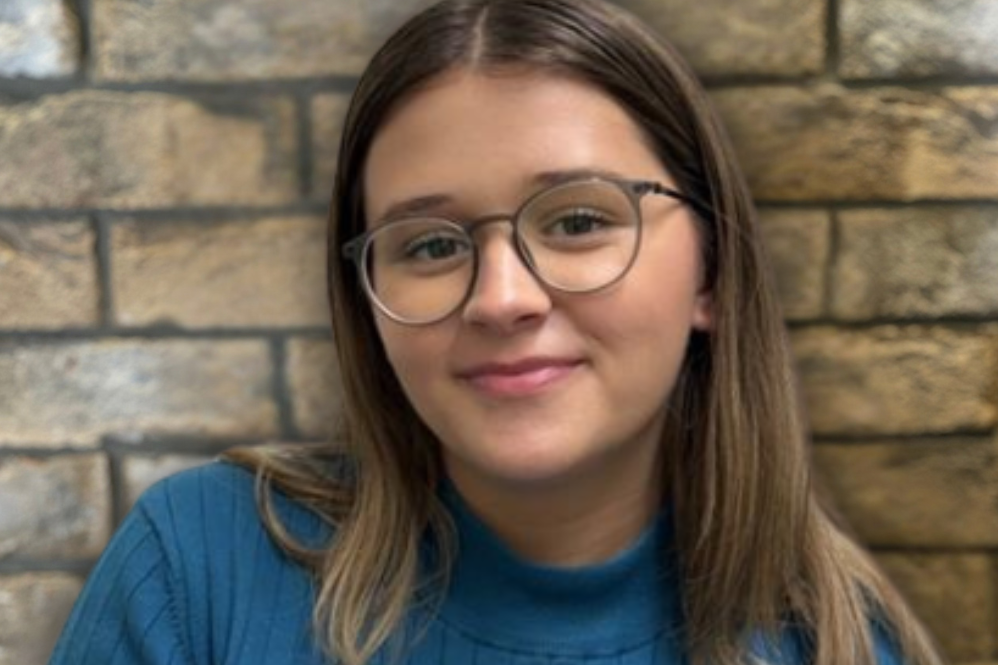 Porträt einer lächelnden jungen Frau mit Brille und blauem Pullover vor einer Steinmauer, die Freundlichkeit und Willkommenskultur ausstrahlt. Porträt einer lächelnden jungen Frau mit Brille und blauem Pullover vor einer Steinmauer, die Freundlichkeit und Willkommenskultur ausstrahlt.