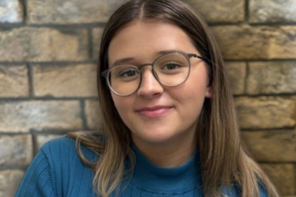 Porträt einer lächelnden jungen Frau mit Brille und blauem Pullover vor einer Steinmauer, die Freundlichkeit und Willkommenskultur ausstrahlt.