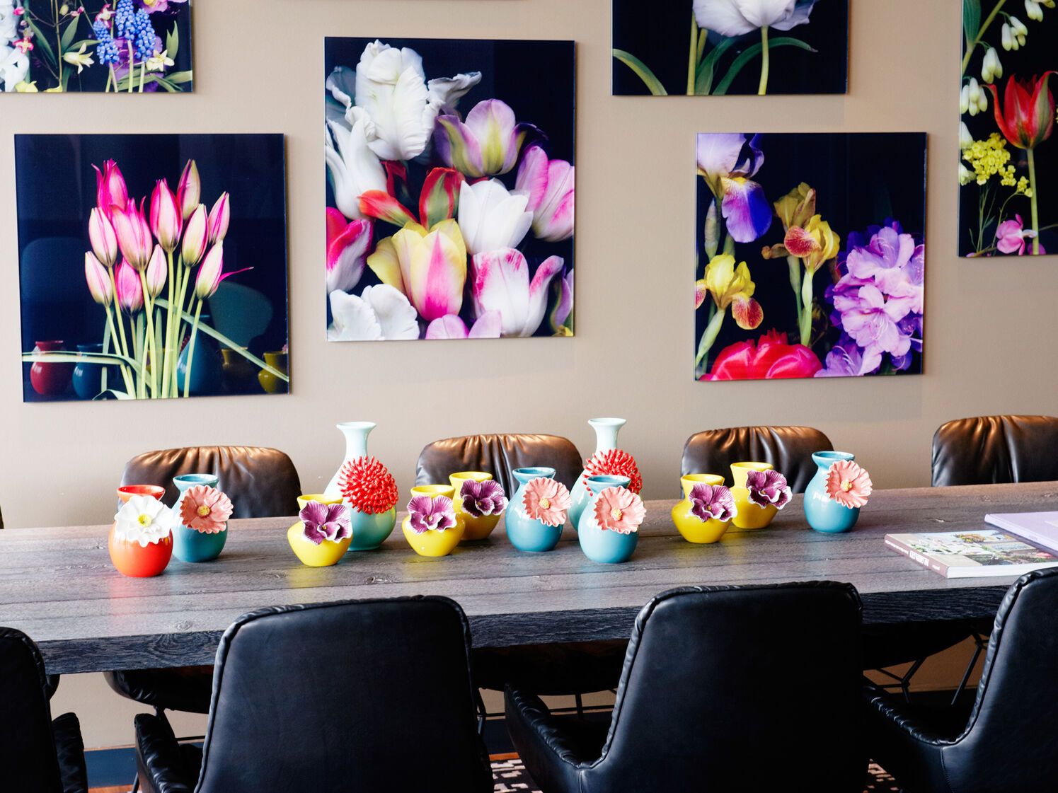 Bunter Konferenztisch mit kleinen, farbenfrohen Vasen und stilisierten Blumenarrangements, umgeben von schwarzen Lederstühlen. Im Hintergrund wandern großformatige, lebendige Blumenbilder an der Wand.