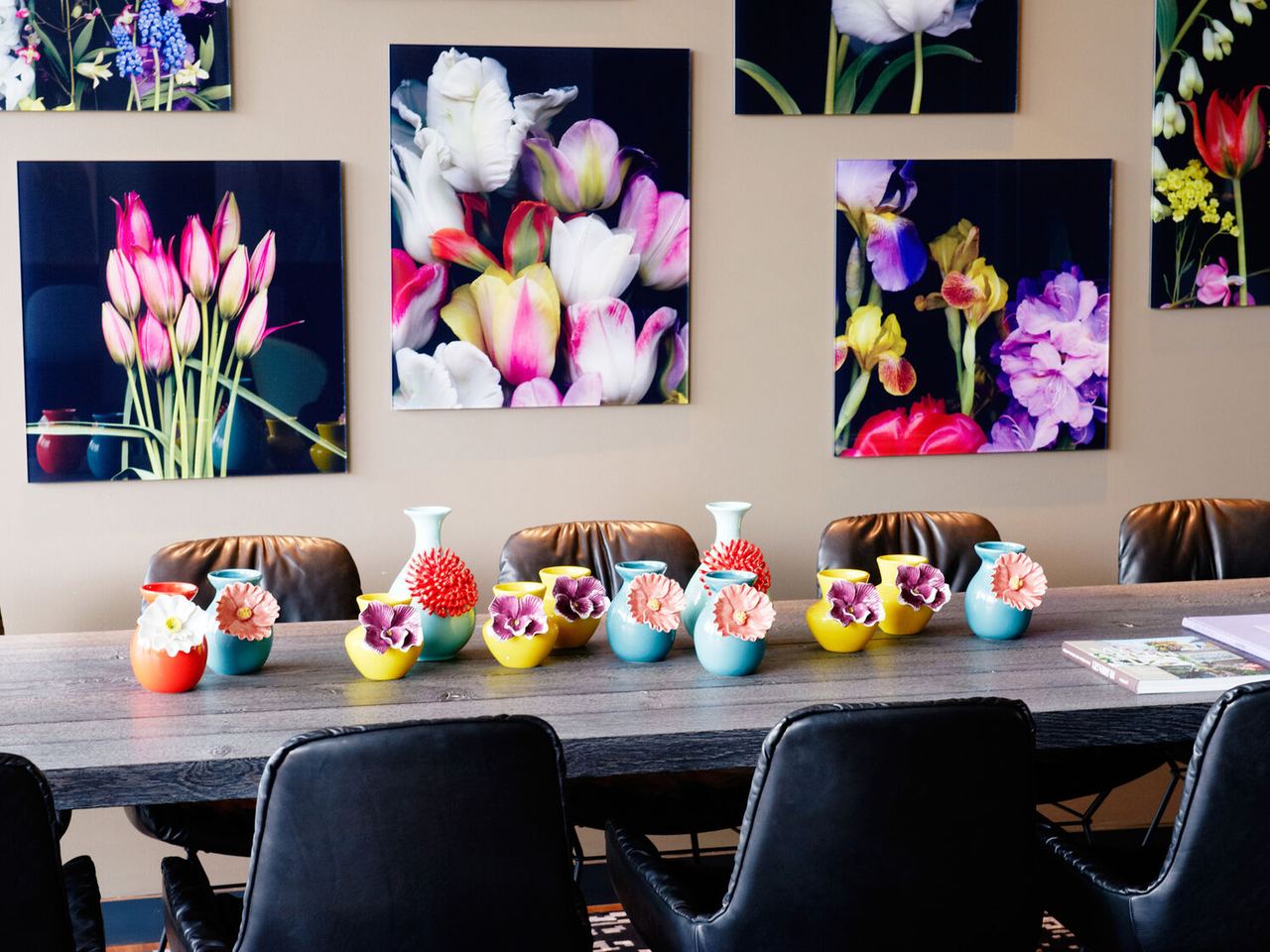 Bunter Konferenztisch mit kleinen, farbenfrohen Vasen und stilisierten Blumenarrangements, umgeben von schwarzen Lederstühlen. Im Hintergrund wandern großformatige, lebendige Blumenbilder an der Wand.