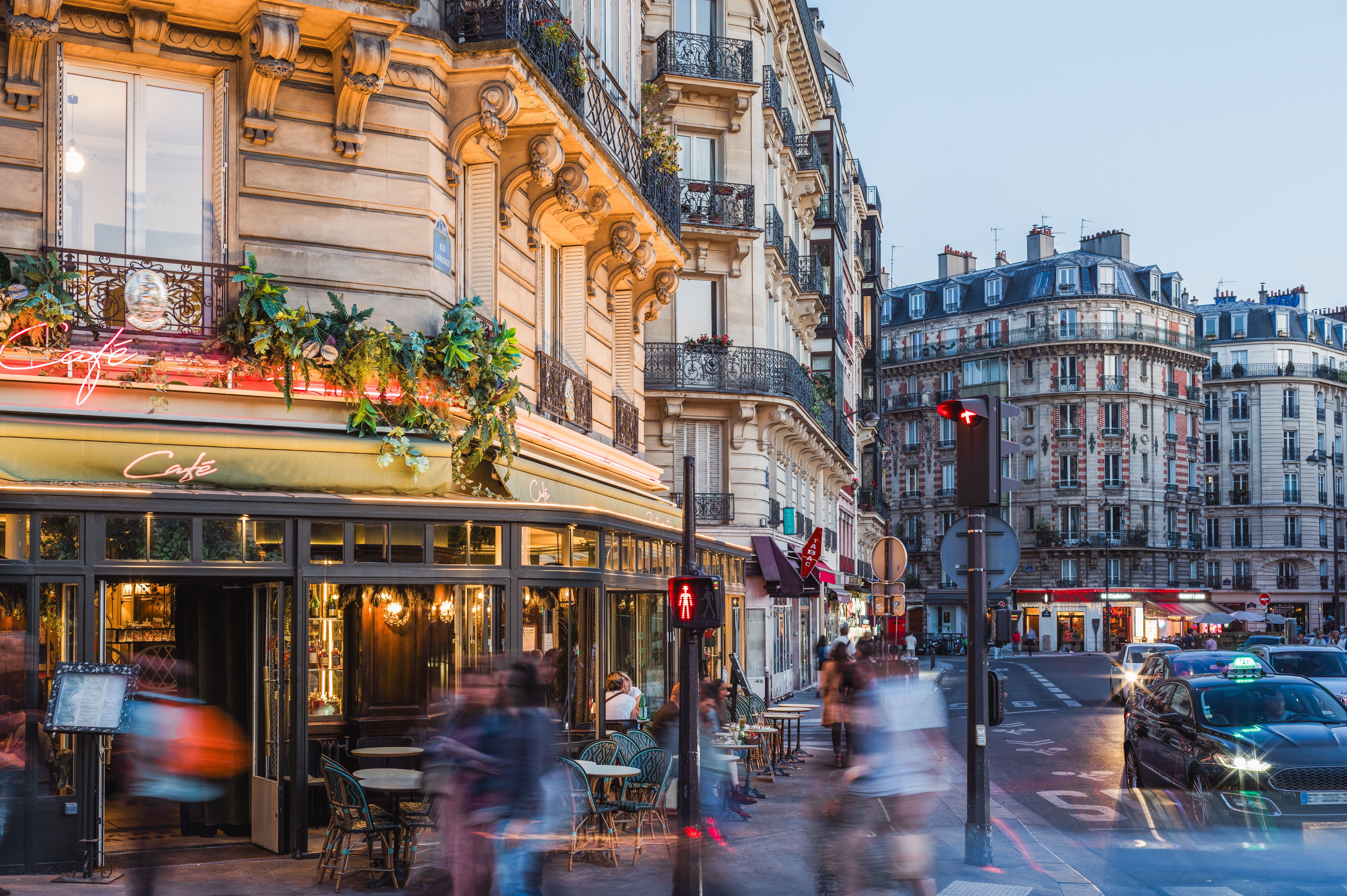 Straßenszene einer belebten Pariser Ecke mit traditionellem Café, Außenbestuhlung und historischen Gebäuden bei Dämmerung, die eine gemütliche und lebendige Atmosphäre vermittelt.