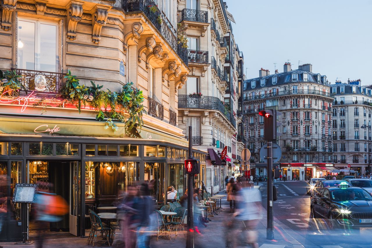 Straßenszene einer belebten Pariser Ecke mit traditionellem Café, Außenbestuhlung und historischen Gebäuden bei Dämmerung, die eine gemütliche und lebendige Atmosphäre vermittelt.