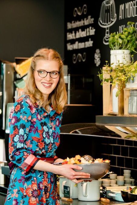 Frau mit Brille und buntem Kleid steht lächelnd an einem Buffet mit frischem Obst und Pflanzen, die Atmosphäre wirkt einladend und gemütlich.