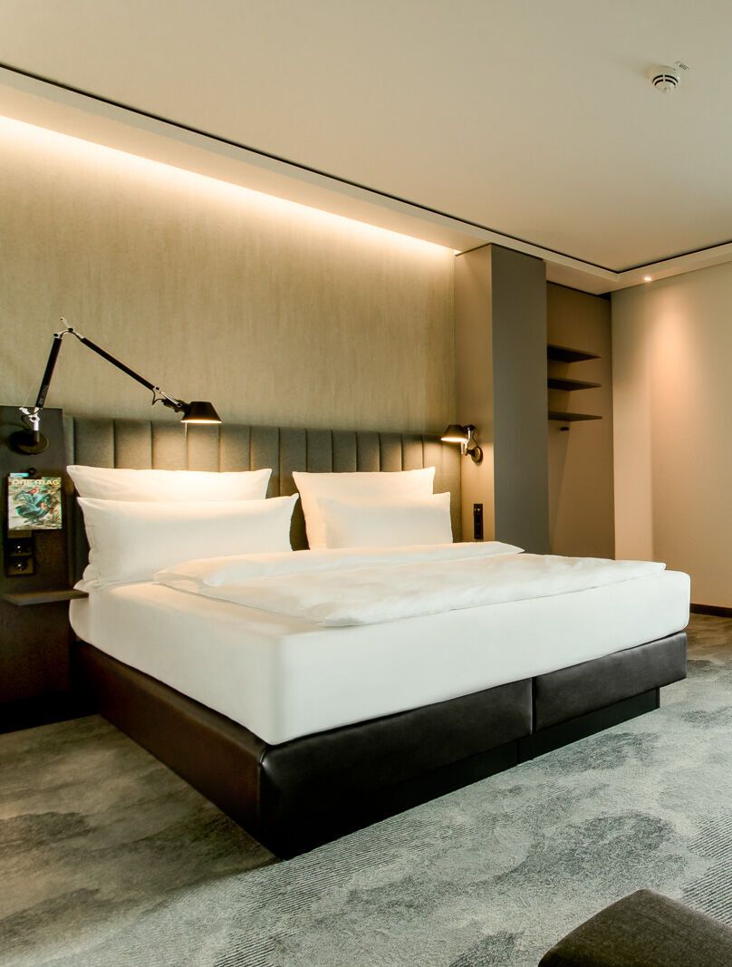 Hotelzimmer mit großem Doppelbett, indirekter Beleuchtung, grünem Vorhang und Blick auf Bäume. Design mit Sitzbereich, Schreibtisch und beruhigender Atmosphäre für erholsamen Aufenthalt.