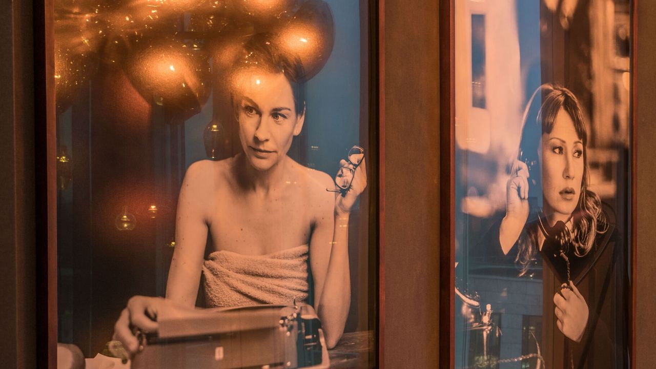 Zwei große gerahmte Fotografien an einer Wand zeigen Frauen in entspannten Momenten: Eine tippt an einer Schreibmaschine, die andere hört mit Kopfhörern Musik. Warme, gemütliche Atmosphäre durch sanfte Beleuchtung.