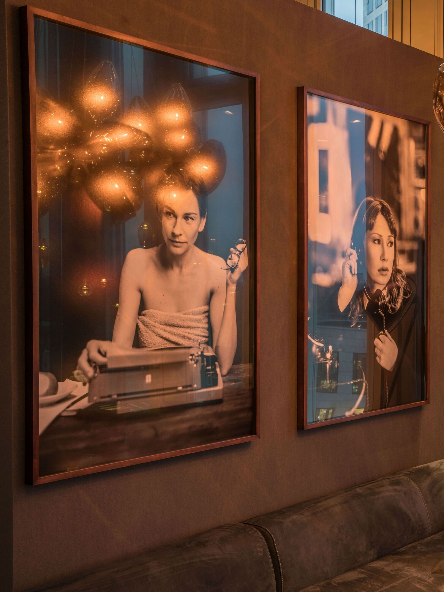 Zwei große gerahmte Fotografien an einer Wand zeigen Frauen in entspannten Momenten: Eine tippt an einer Schreibmaschine, die andere hört mit Kopfhörern Musik. Warme, gemütliche Atmosphäre durch sanfte Beleuchtung.