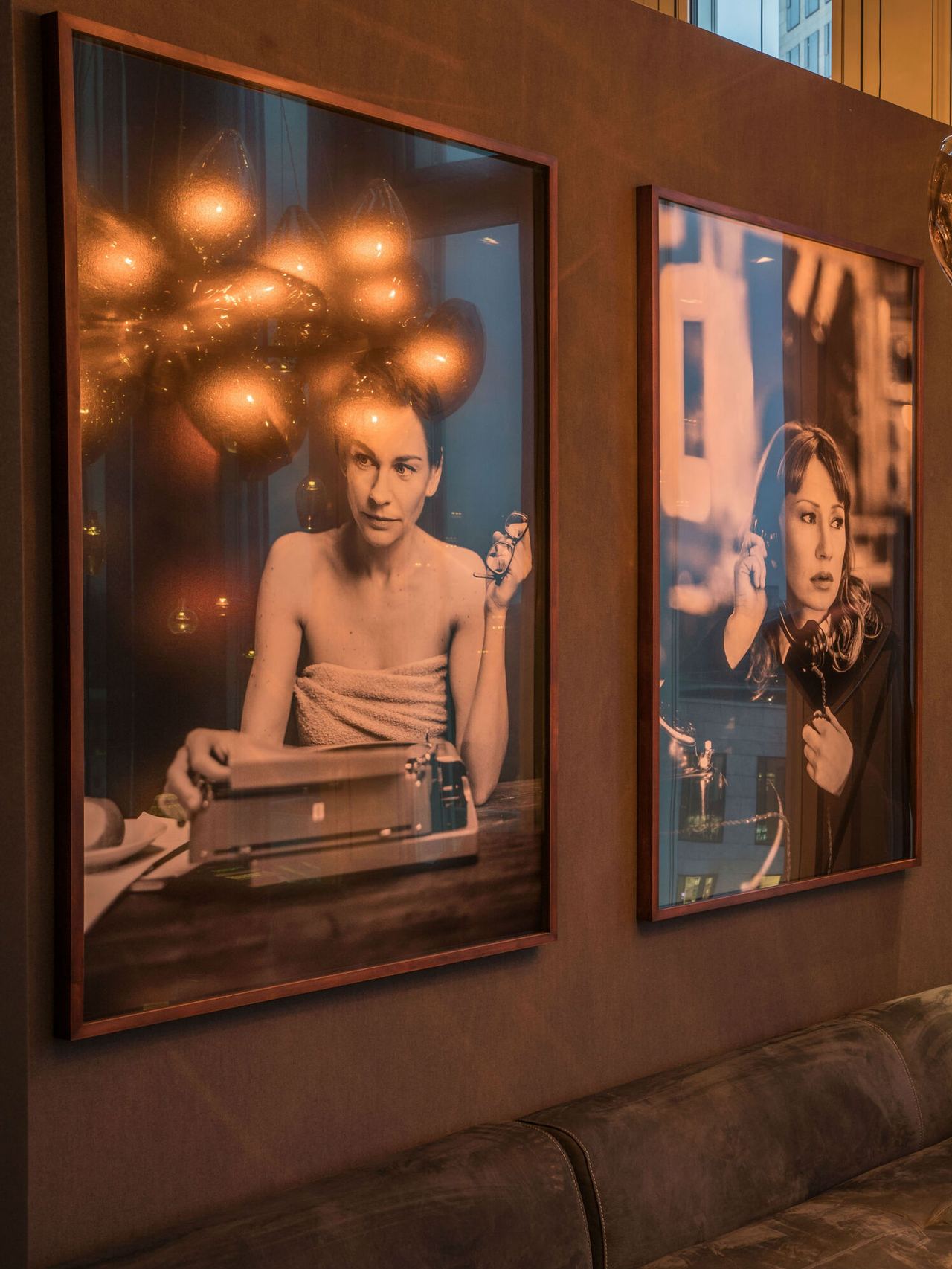 Zwei große gerahmte Fotografien an einer Wand zeigen Frauen in entspannten Momenten: Eine tippt an einer Schreibmaschine, die andere hört mit Kopfhörern Musik. Warme, gemütliche Atmosphäre durch sanfte Beleuchtung.