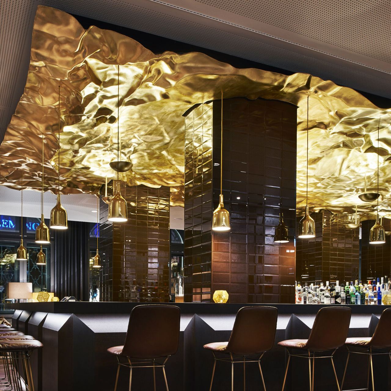 Moderne Hotelbar mit dunklen Sitzgelegenheiten und einer markanten, gold glänzenden Decke, die ein warmes, luxuriöses Ambiente schafft. Sanfte Pendelleuchten betonen den eleganten Bartresen.