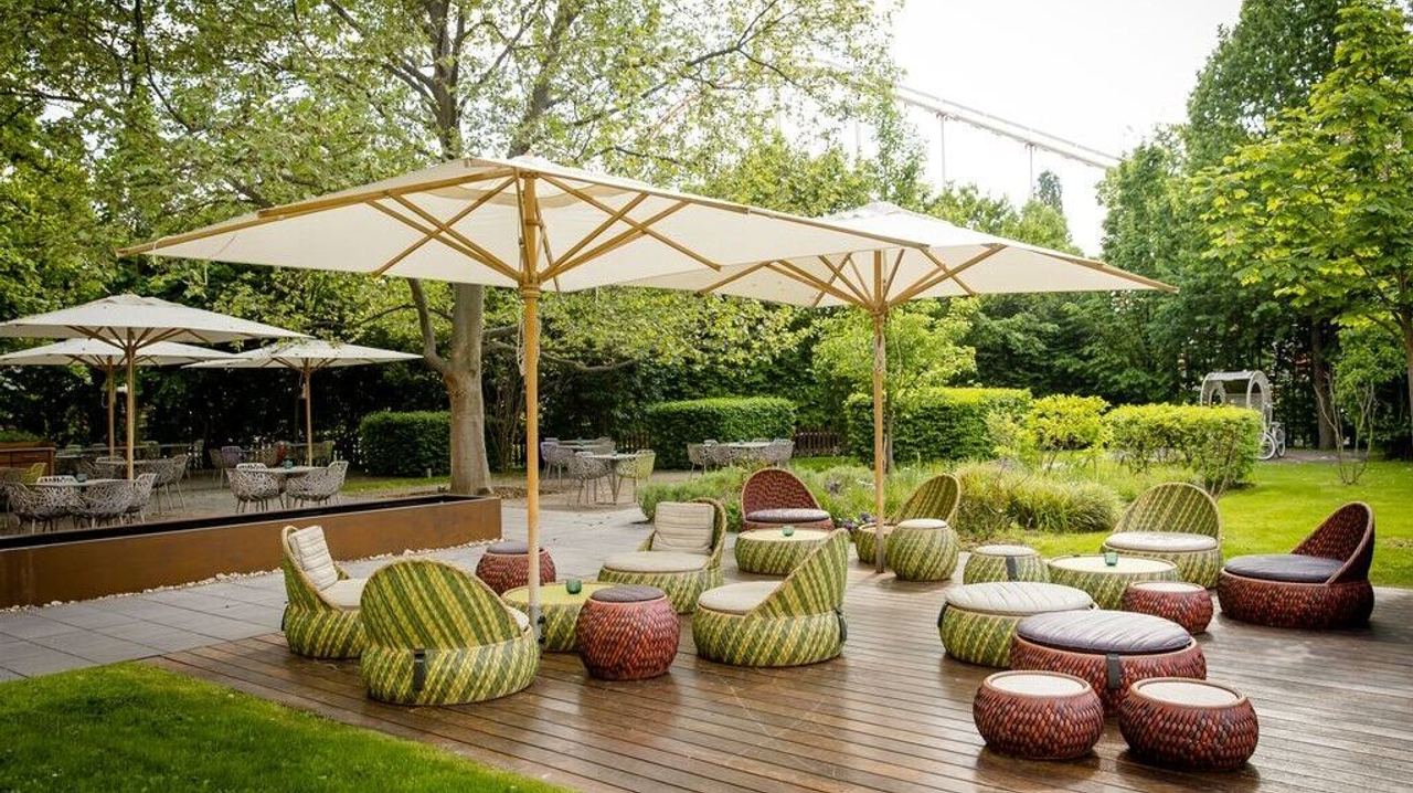 Gemütliche Außensitzplätze mit runden Sitzmöbeln und großen Sonnenschirmen auf einer Holzterrasse, umgeben von grüner, gepflegter Gartenanlage für entspannte Momente im Freien.