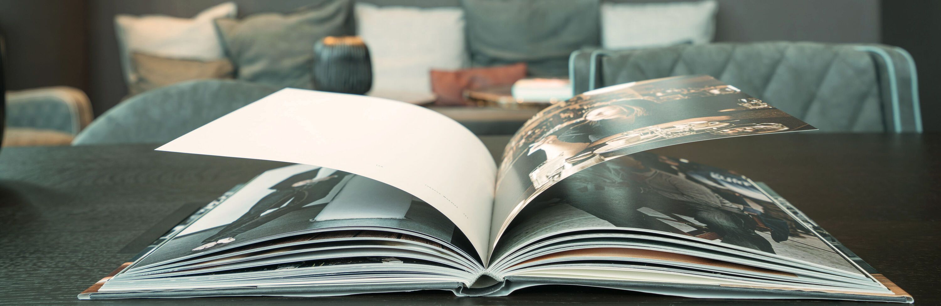 Offenes Fotobuch auf einem Tisch, das Bilder in Schwarz-Weiß zeigt, umgeben von einem gemütlichen, stilvollen Wohnbereich mit Sofas und Kissen im Hintergrund.