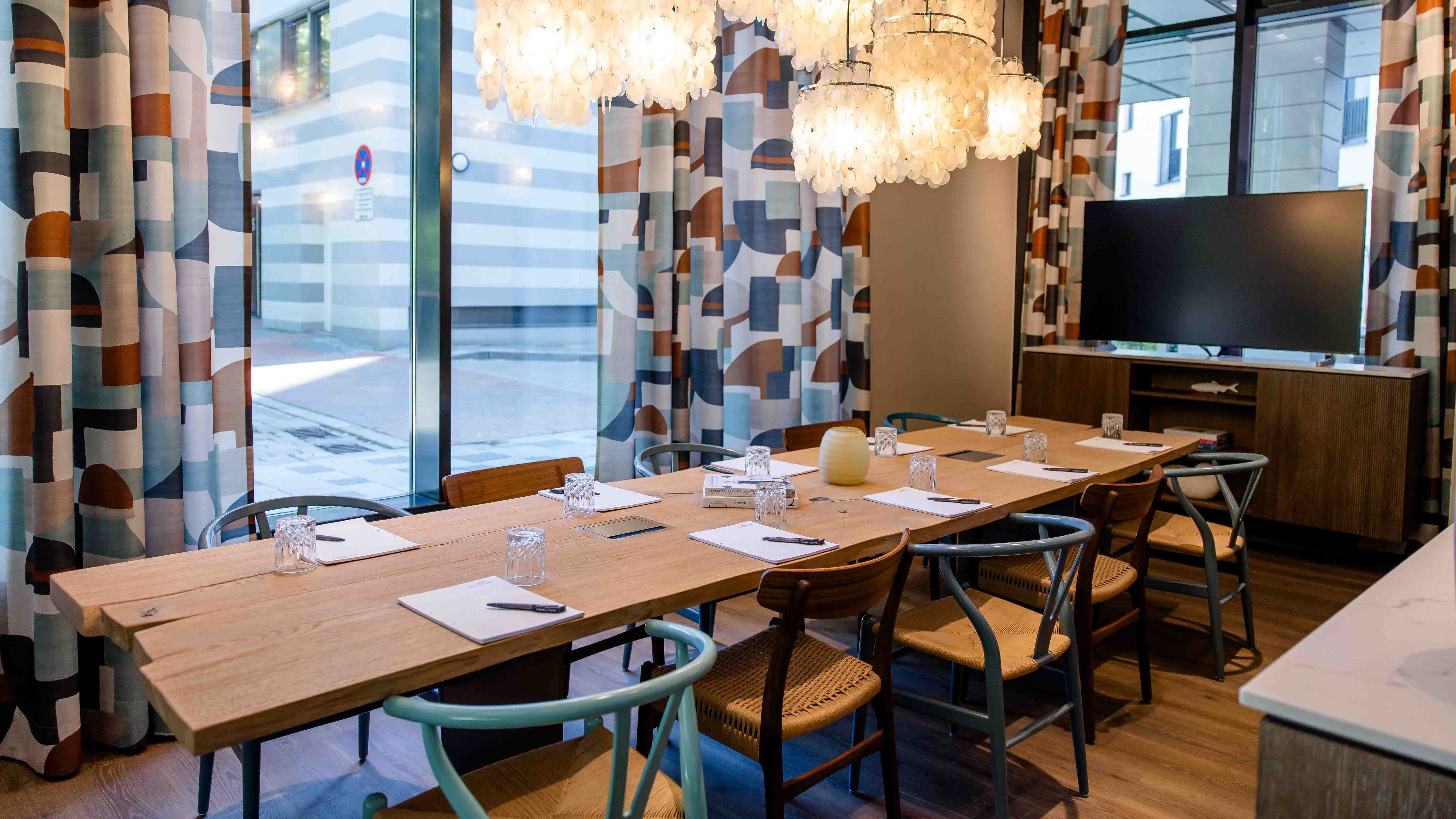 Motel One Konferenzraum mit großem Holztisch, bequemen Stühlen und stilvollen, leuchtenden Lampen. Große Fenster bieten Tageslicht und Blick auf die Stadt. Ideal für Besprechungen oder Gruppenarbeiten.