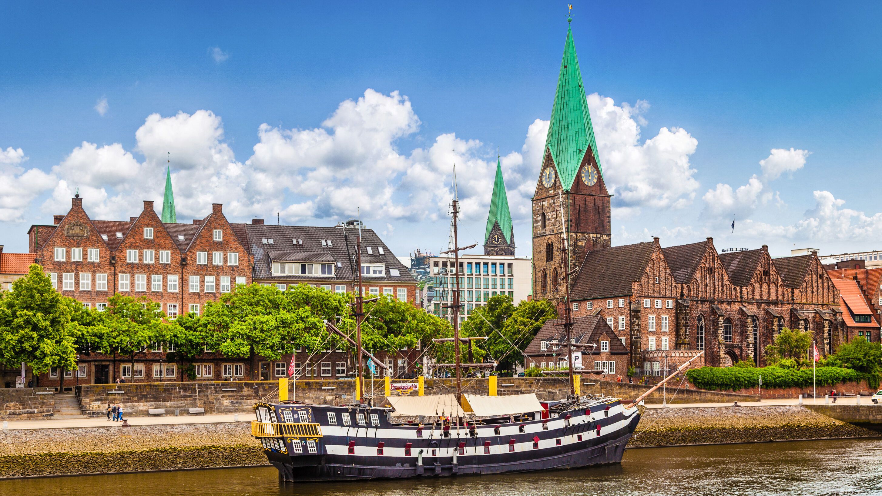 Blick auf eine historische Stadt mit roten Backsteingebäuden und hohen, grünen Kirchtürmen am Flussufer. Ein traditionelles Segelschiff liegt am Ufer, während in der Flussmitte ein Ruderboot mit vier Personen unterwegs ist. Der Himmel ist blau mit vereinzelten Wolken.